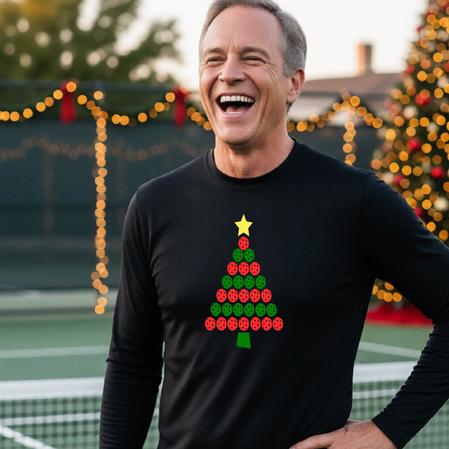 A smiling man wearing a black long-sleeve shirt with a Christmas tree made of red and green pickleballs and a yellow star, standing on a pickleball court with holiday lights in the background.