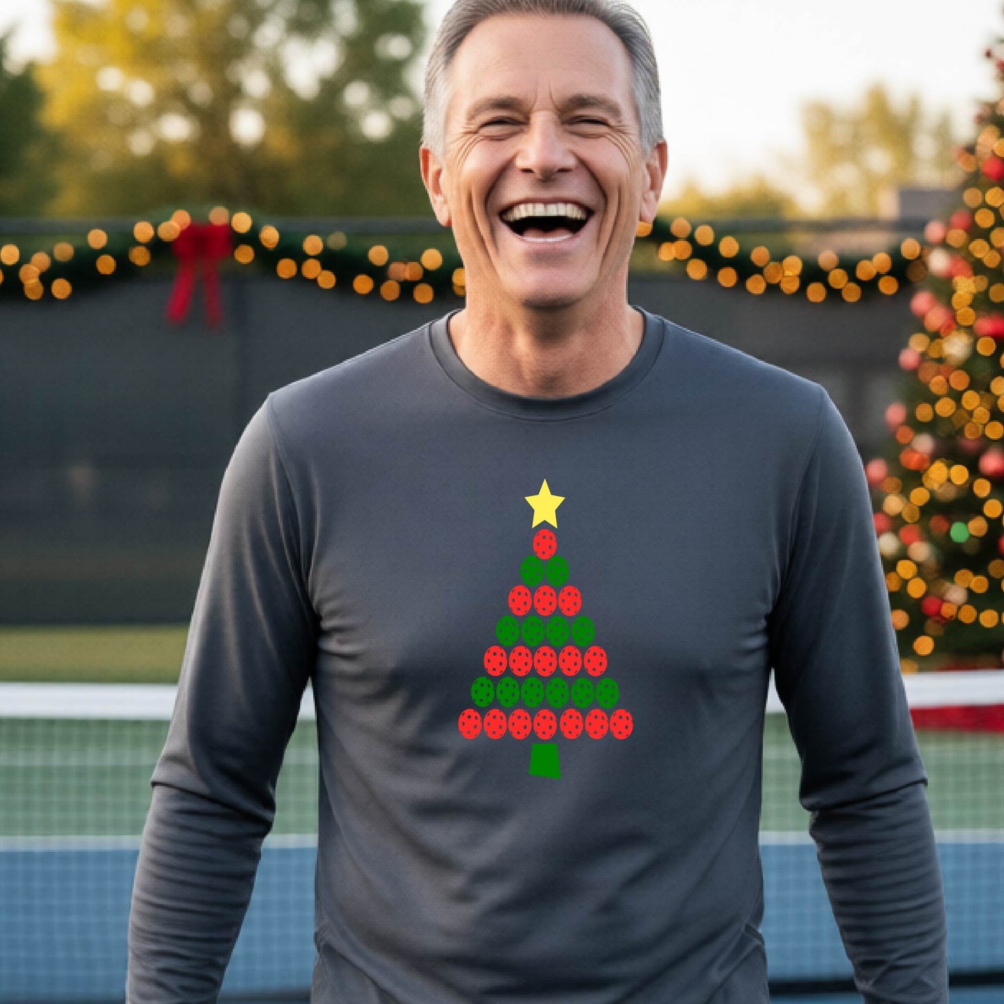 A smiling man wearing a dark gray long-sleeve shirt with a Christmas tree made of red and green pickleballs and a yellow star, standing on a pickleball court with holiday lights in the background.