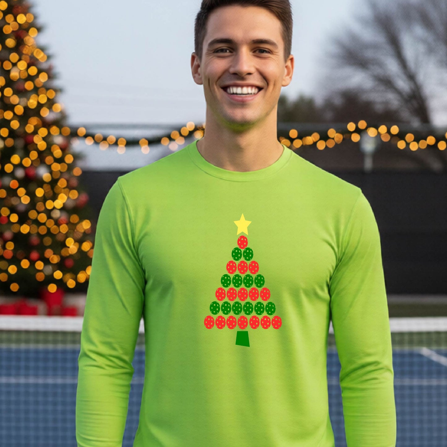 A smiling man wearing a lime green long-sleeve shirt with a Christmas tree made of red and green pickleballs and a yellow star, standing on a pickleball court with holiday lights in the background.