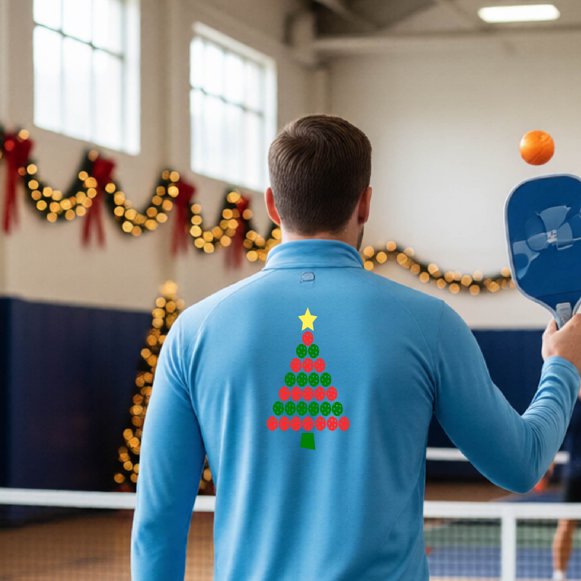 A smiling man wearing a light blue long-quarter zip sleeve shirt with a Christmas tree made of red and green pickleballs and a yellow star, standing on a pickleball court with holiday lights in the background.