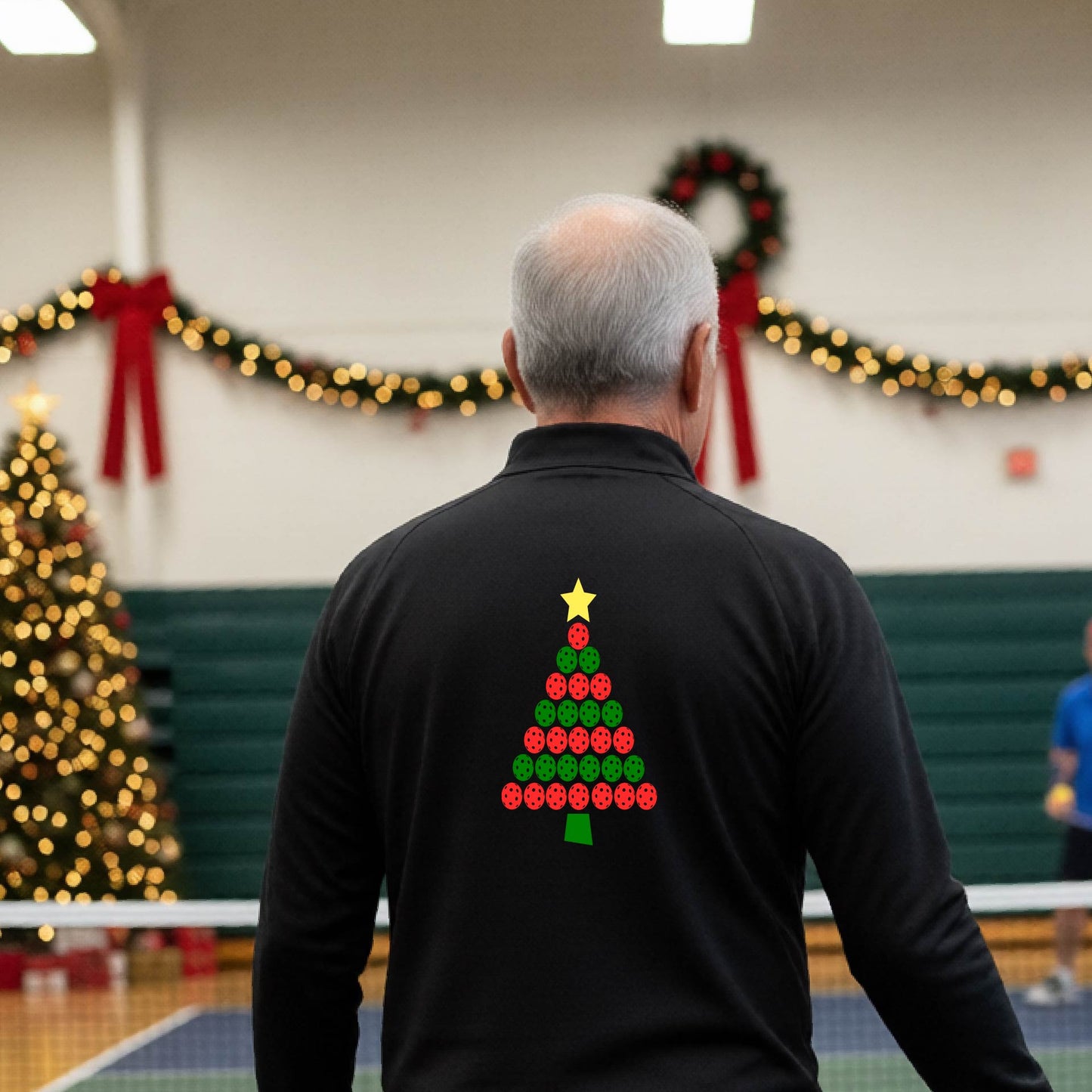 A smiling man wearing a black long-quarter zip sleeve shirt with a Christmas tree made of red and green pickleballs and a yellow star, standing on a pickleball court with holiday lights in the background.