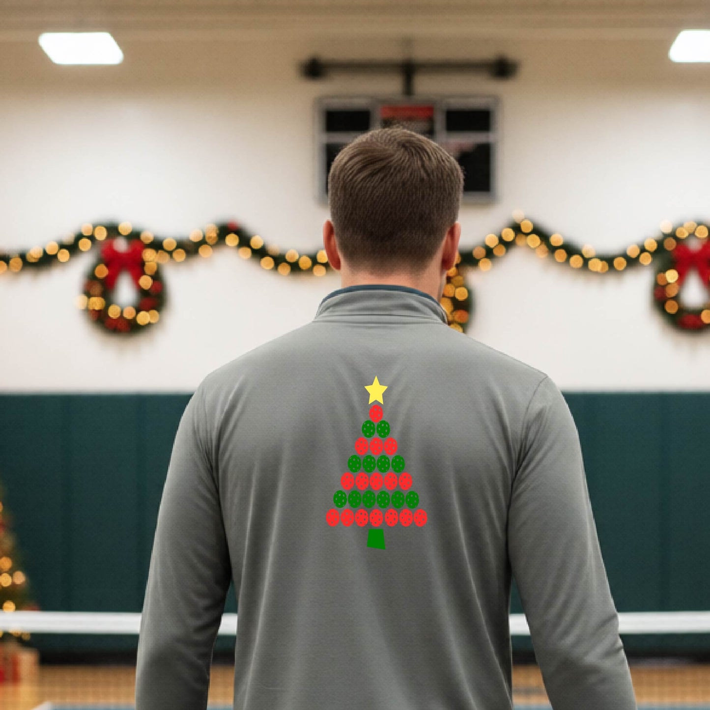A smiling man wearing a gray long-quarter zip sleeve shirt with a Christmas tree made of red and green pickleballs and a yellow star, standing on a pickleball court with holiday lights in the background.
