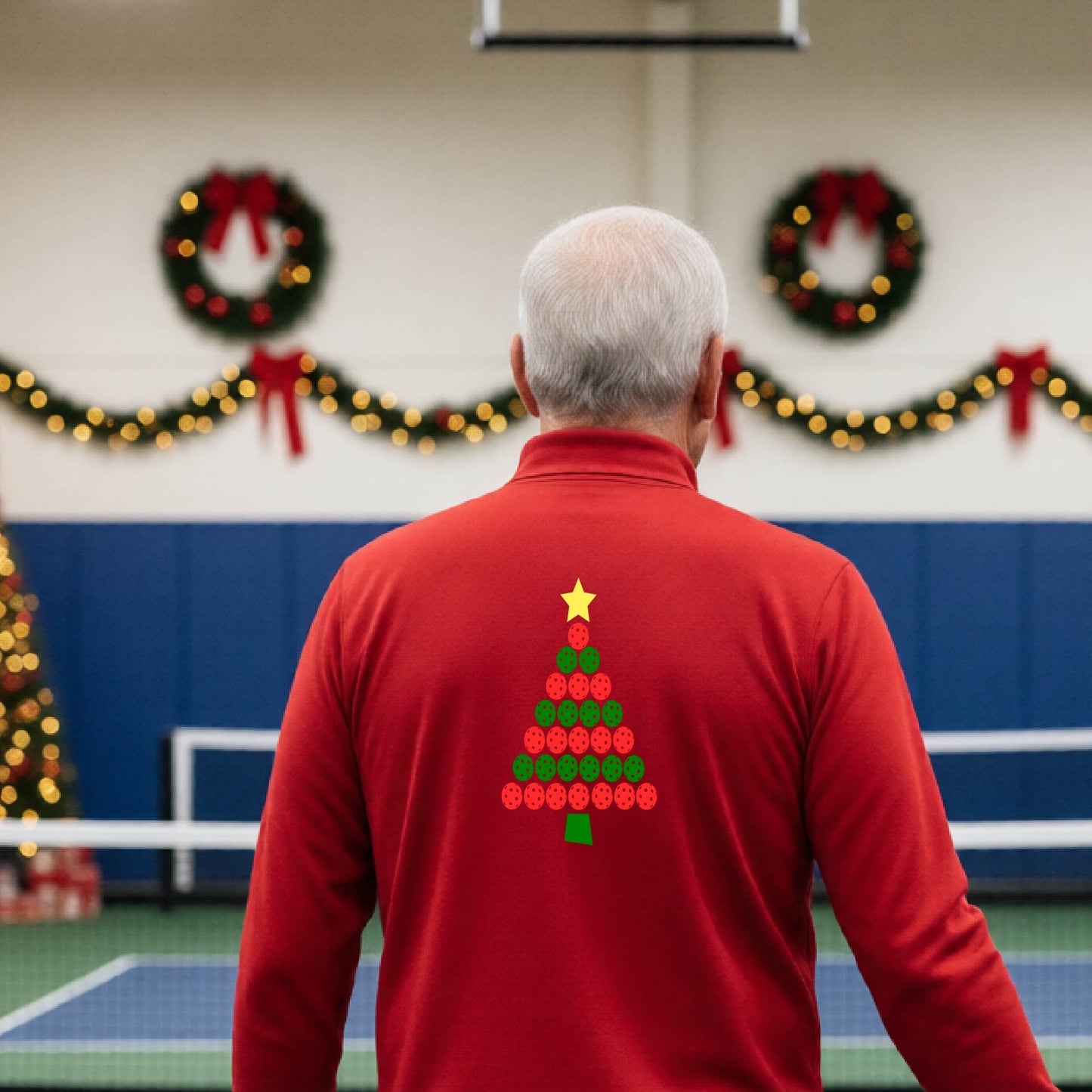 A smiling man wearing a red long-quarter zip sleeve shirt with a Christmas tree made of red and green pickleballs and a yellow star, standing on a pickleball court with holiday lights in the background.