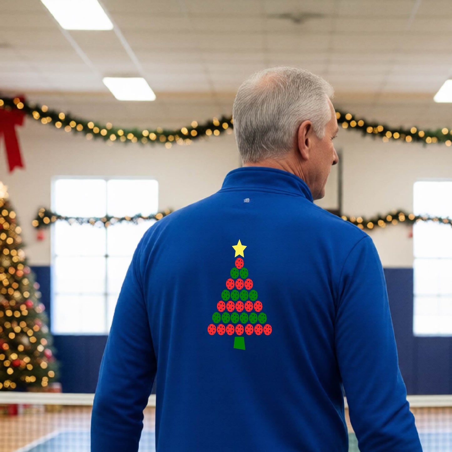 A smiling man wearing a royal blue long-quarter zip sleeve shirt with a Christmas tree made of red and green pickleballs and a yellow star, standing on a pickleball court with holiday lights in the background.