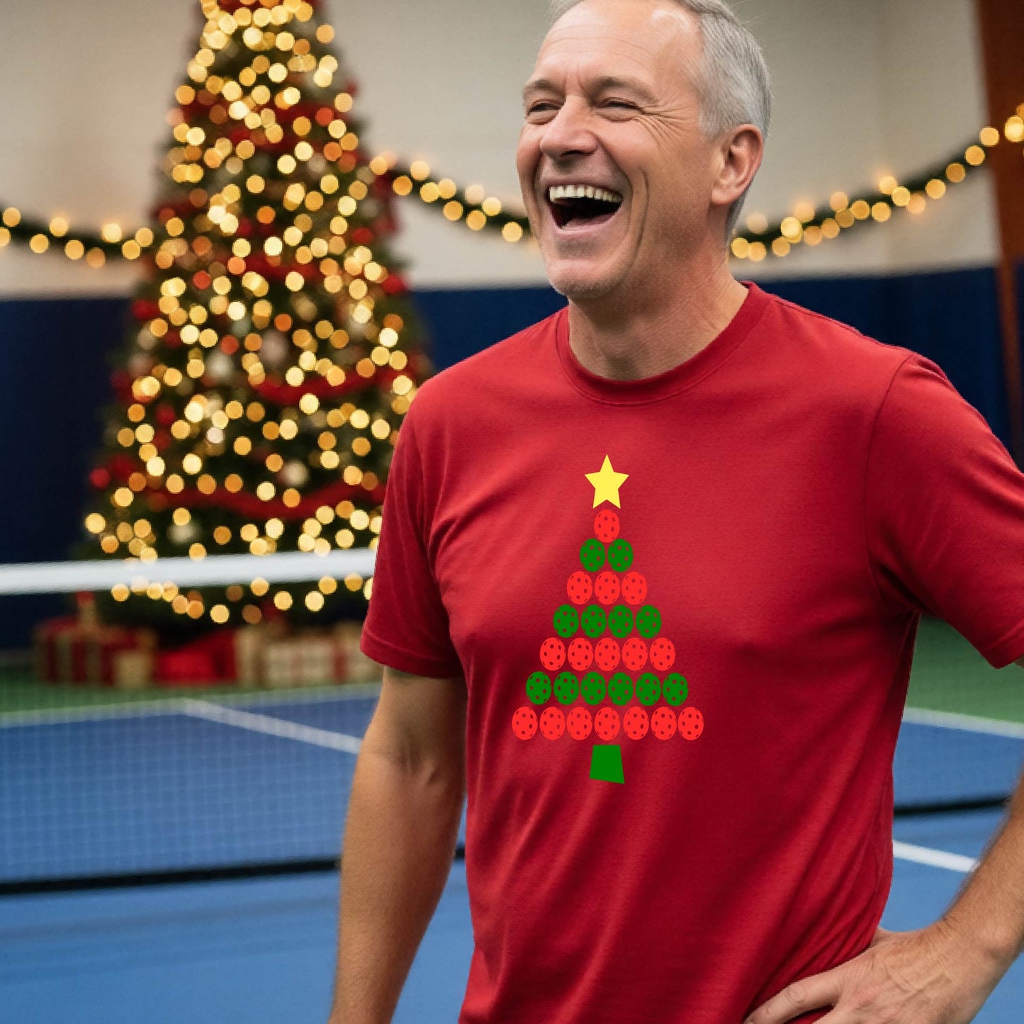 A smiling man wearing a red short-sleeve t-shirt with a Christmas tree made of pickleball balls, standing on a pickleball court with Christmas light and decoration in the background. 