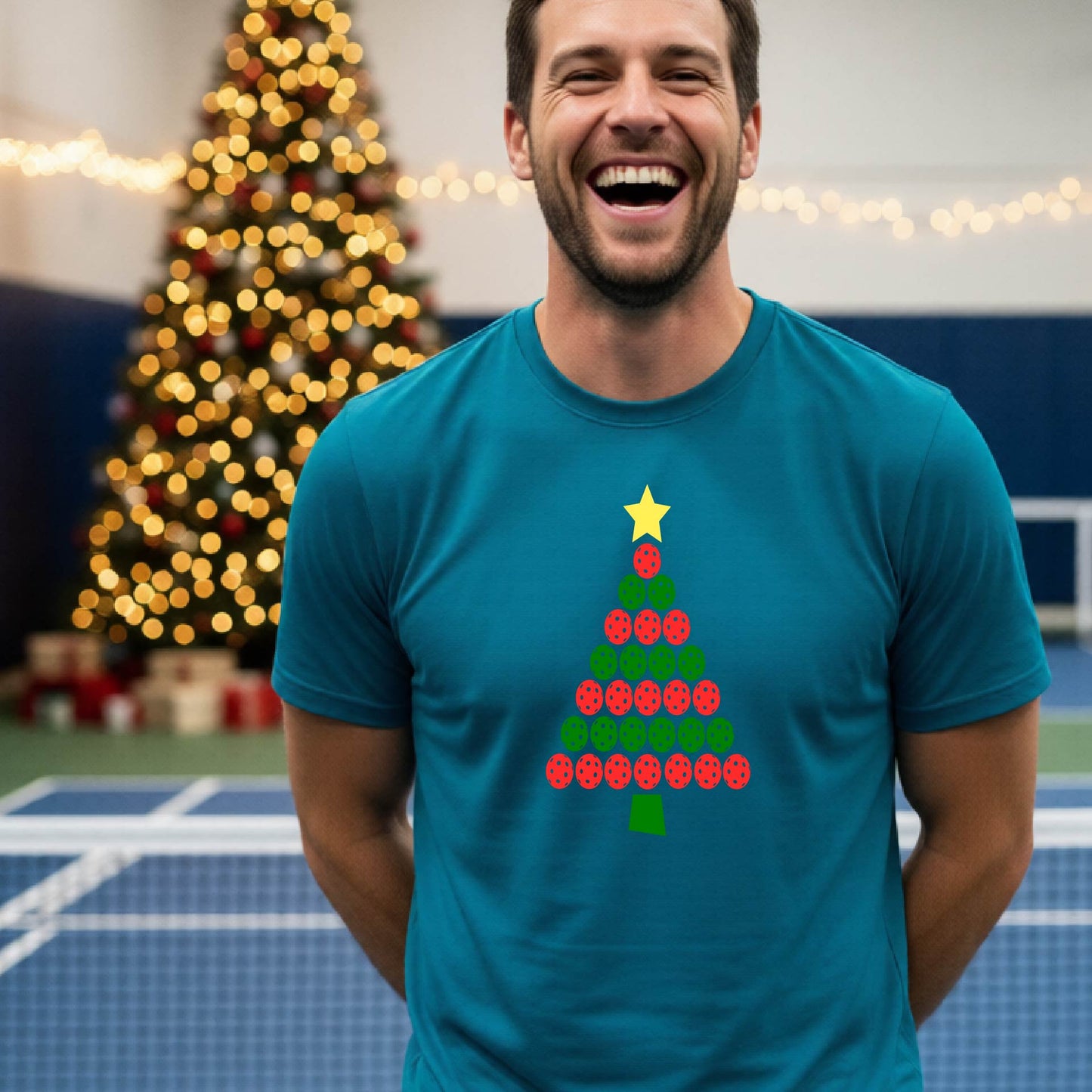 A smiling man wearing a blue short-sleeve t-shirt with a Christmas tree made of pickleball balls, standing on a pickleball court with Christmas light and decoration in the background. 