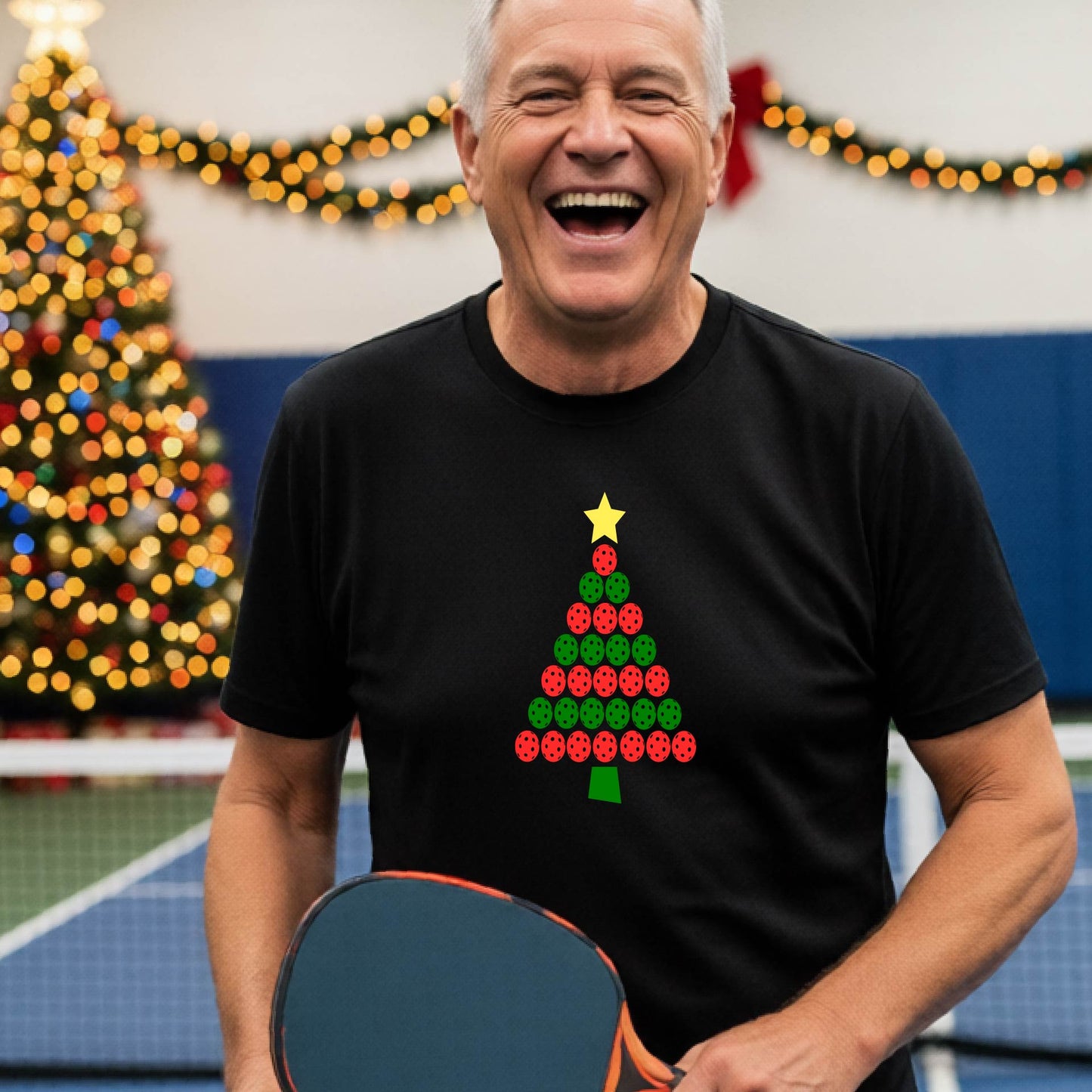 A smiling man wearing a black short-sleeve t-shirt with a Christmas tree made of pickleball balls, standing on a pickleball court with Christmas light and decoration in the background. 
