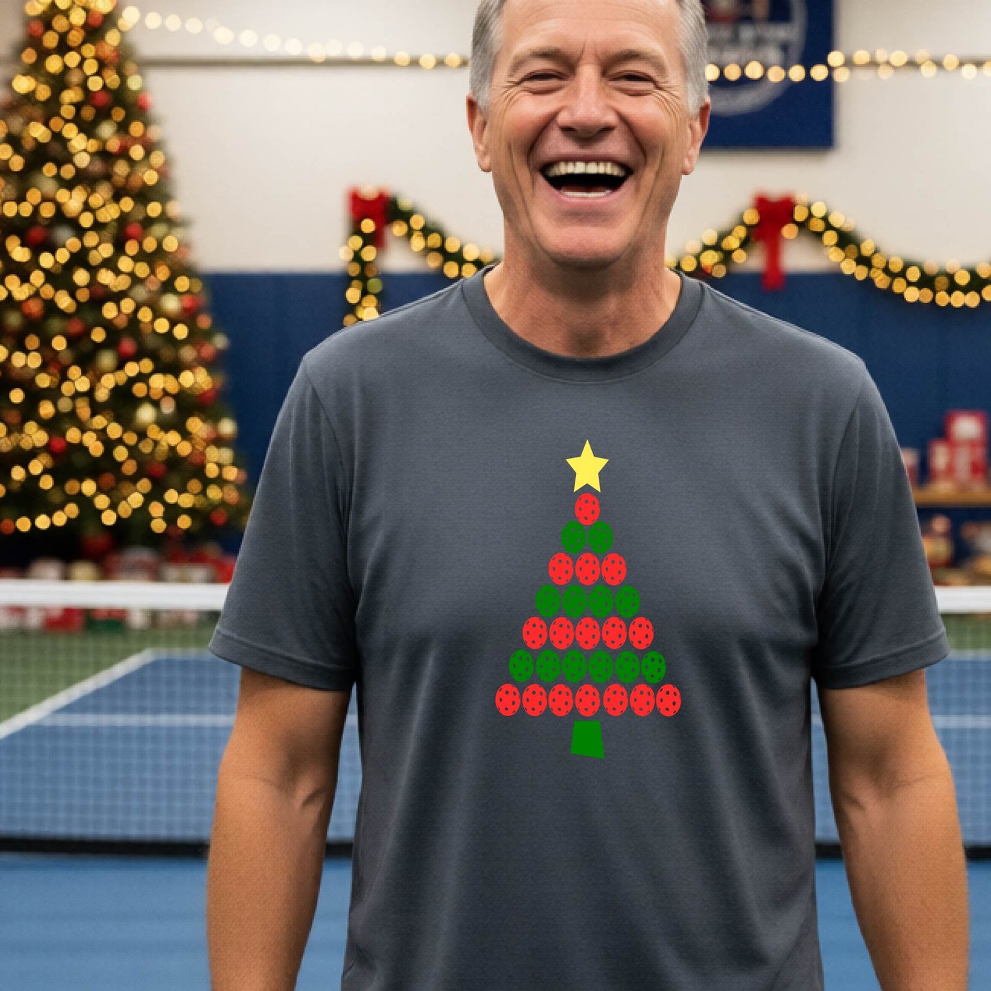 A smiling man wearing a gray short-sleeve t-shirt with a Christmas tree made of pickleball balls, standing on a pickleball court with Christmas light and decoration in the background. 