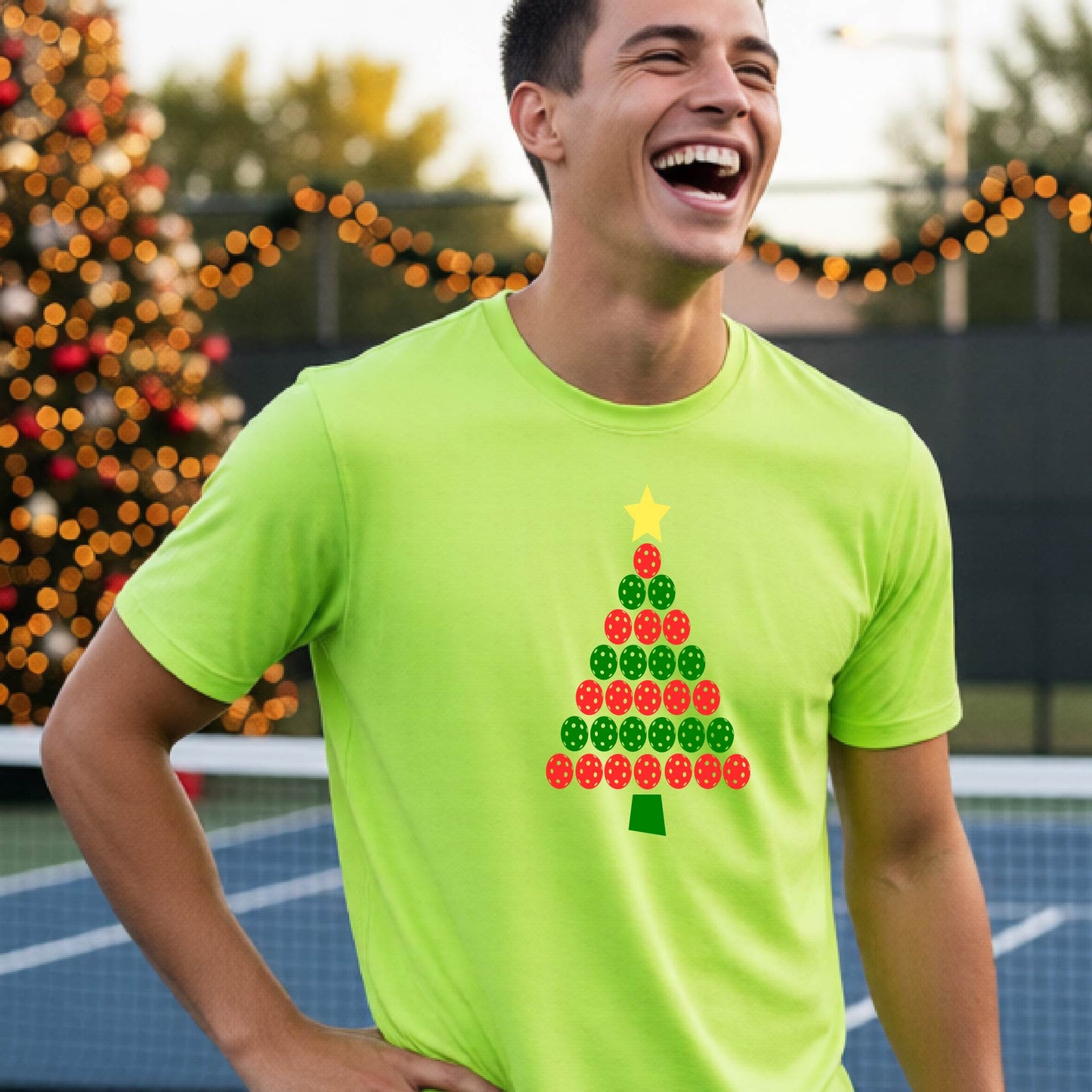 A smiling man wearing a lime green short-sleeve t-shirt with a Christmas tree made of pickleball balls, standing on a pickleball court with Christmas light and decoration in the background. 