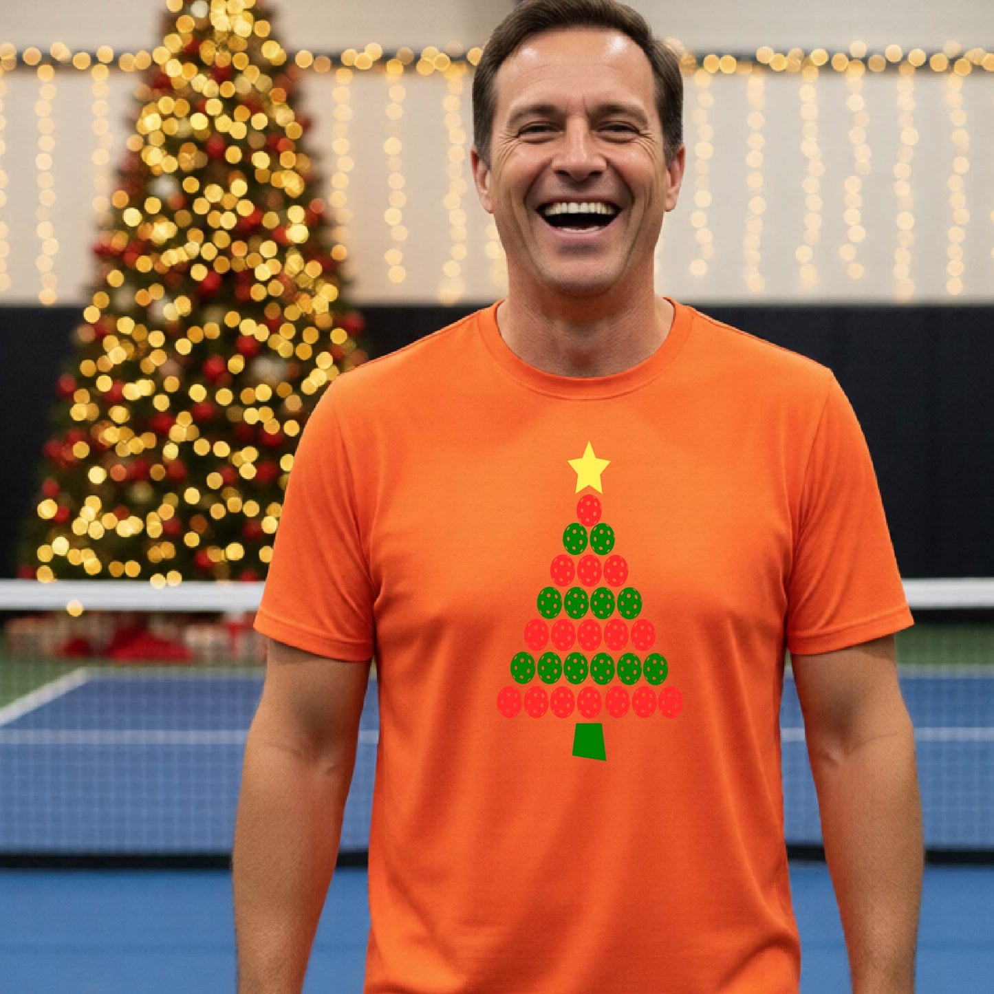 A smiling man wearing an orange short-sleeve t-shirt with a Christmas tree made of pickleball balls, standing on a pickleball court with Christmas light and decoration in the background. 
