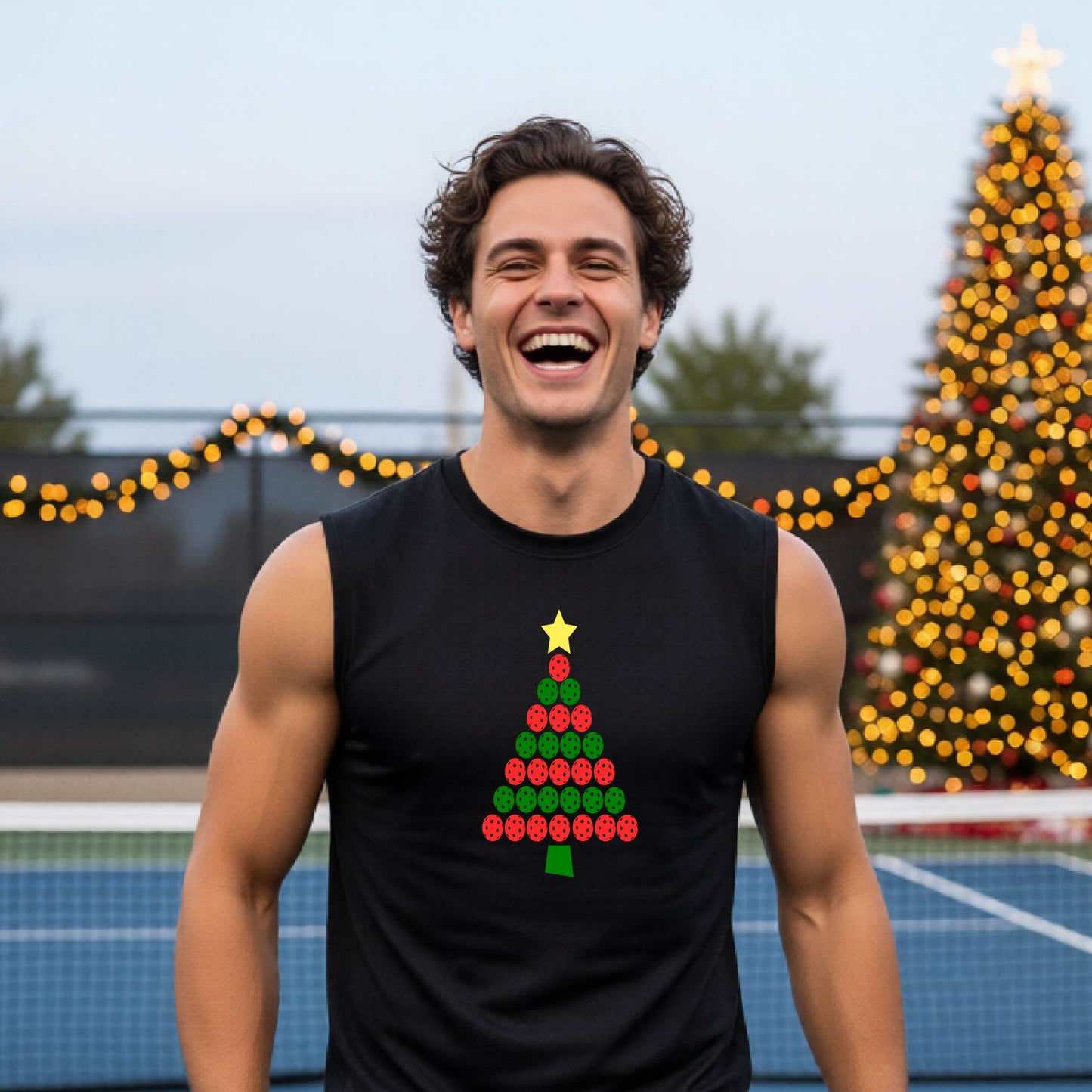 A smiling man wearing a black sleeveless shirt with a Christmas tree made of red and green pickleballs and a yellow star, standing on a pickleball court with holiday lights in the background.