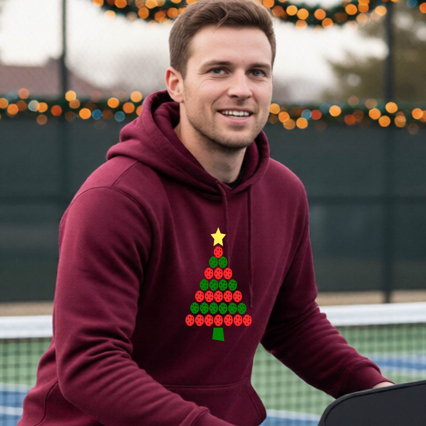 A man wearing a maroon hoodie with a Christmas tree made of red and green pickleballs and a yellow star, in front of a pickleball court.