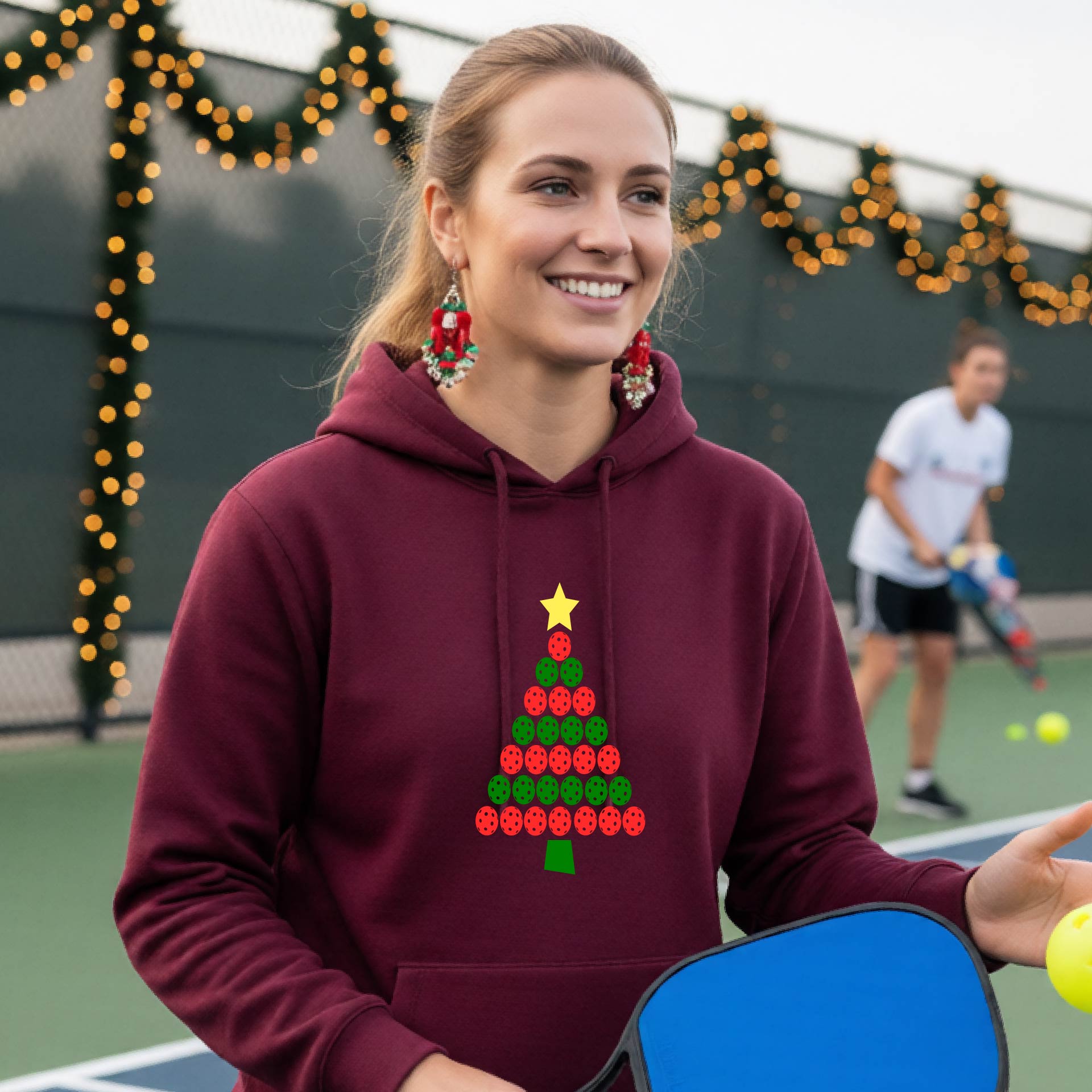 A woman wearing a maroon hoodie with a Christmas tree made of red and green pickleballs and a yellow star, in front of a pickleball court.