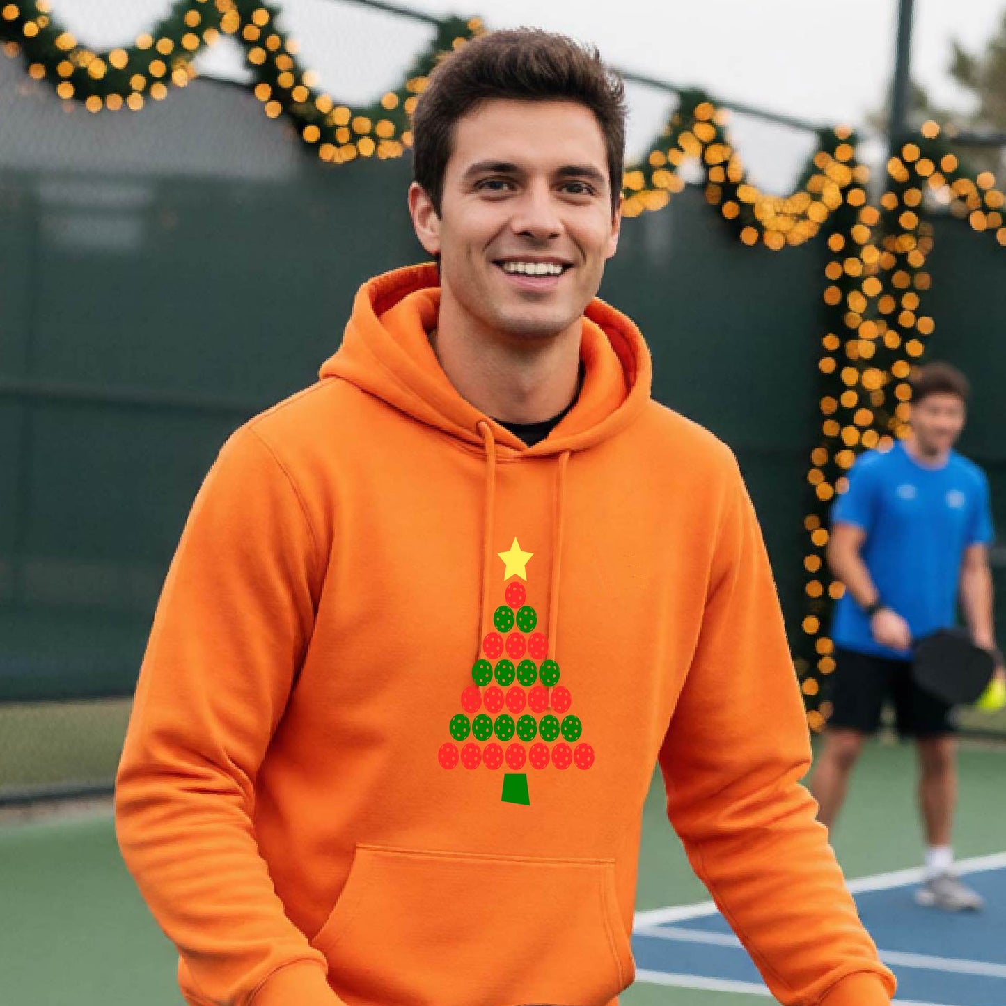 A man wearing a bright orange hoodie with a Christmas tree made of red and green pickleballs and a yellow star, in front of a pickleball court.