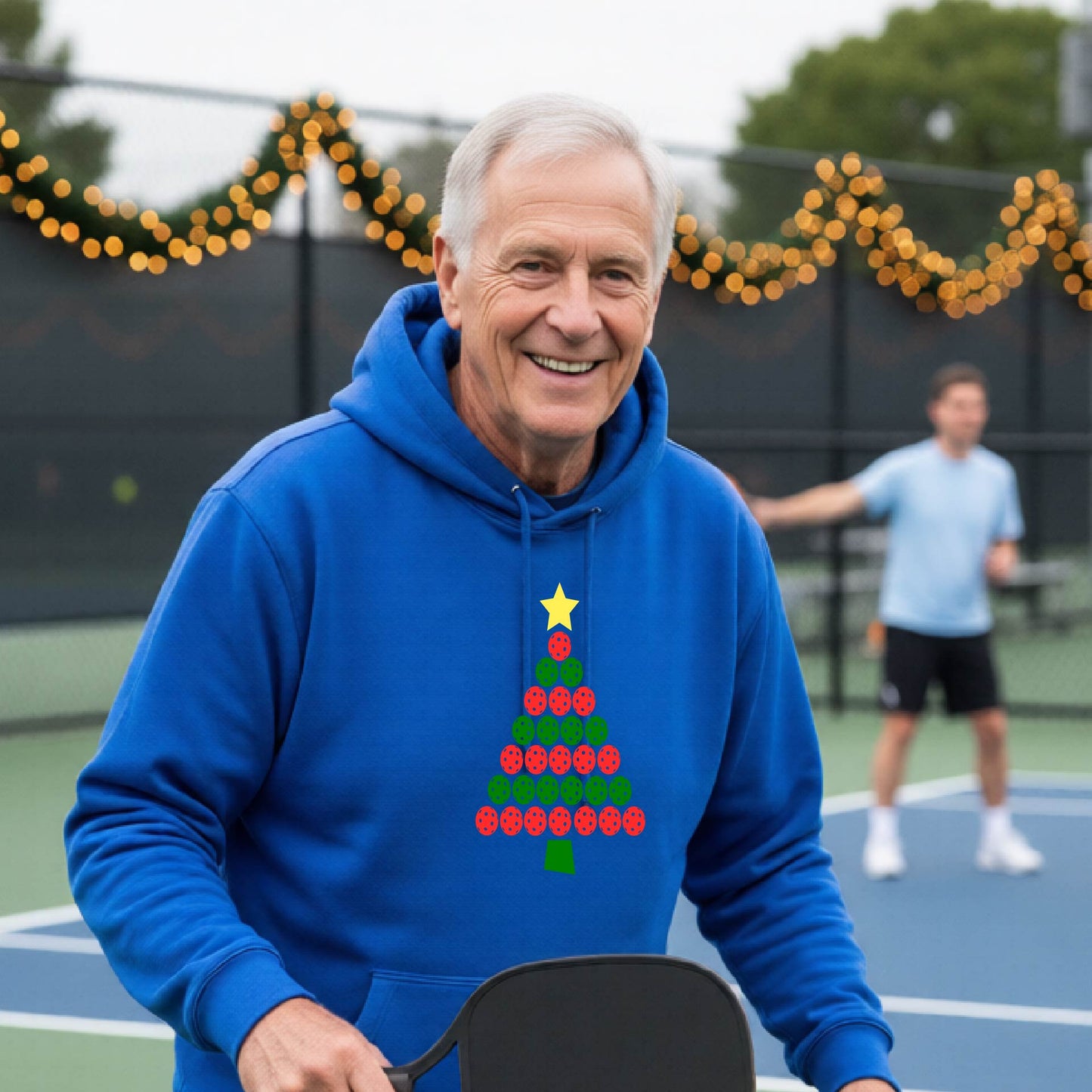 A man wearing a royal blue hoodie with a Christmas tree made of red and green pickleballs and a yellow star, in front of a pickleball court.