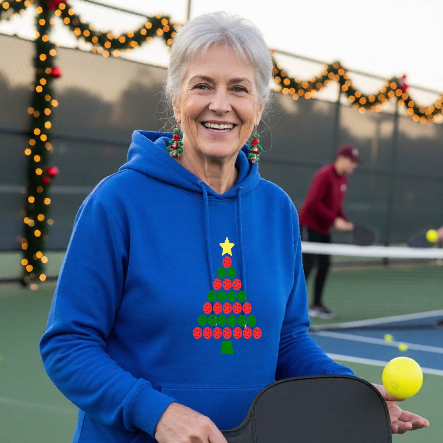 A woman wearing a bright royal blue hoodie with a Christmas tree made of red and green pickleballs and a yellow star, in front of a pickleball court.