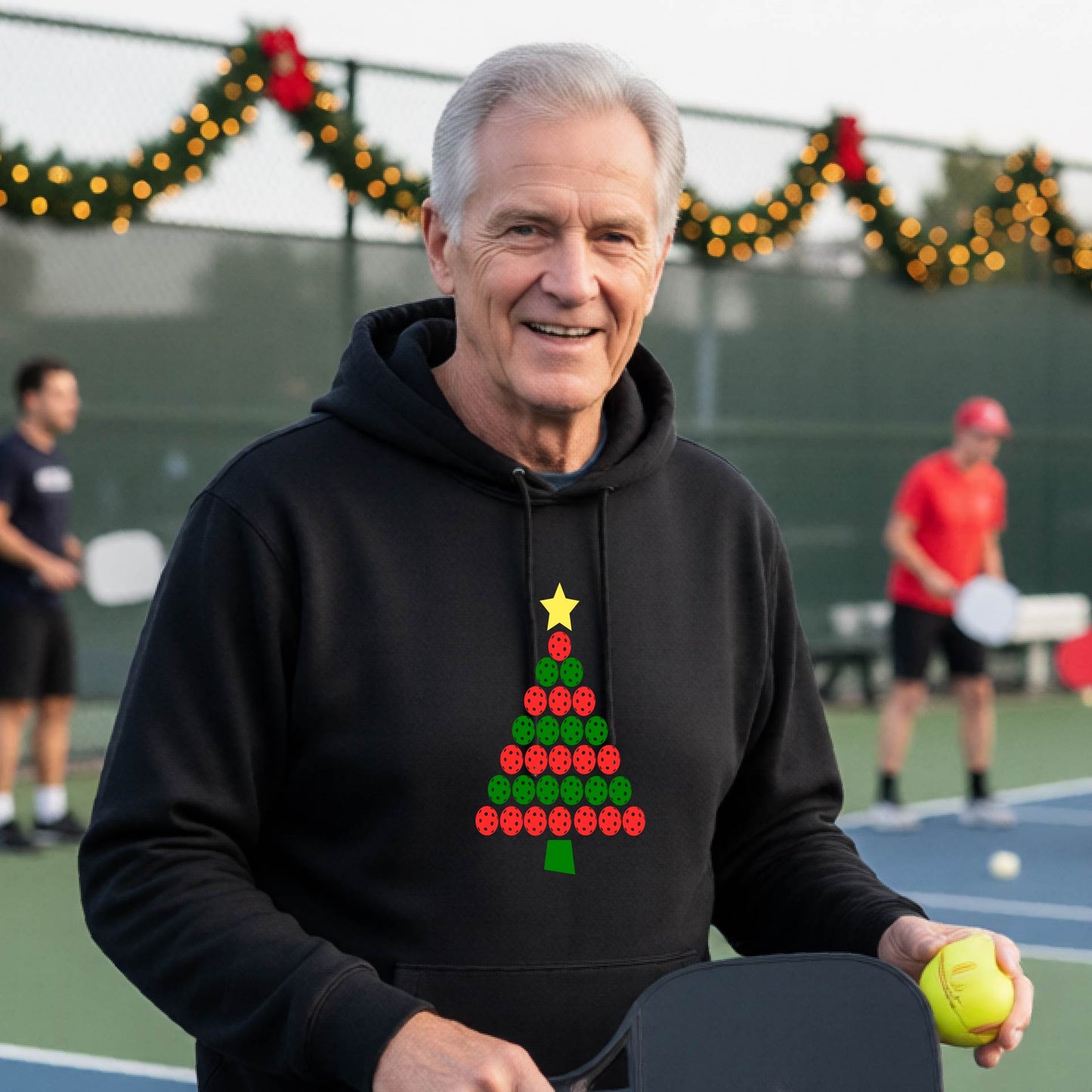 A man wearing a black hoodie with a Christmas tree made of red and green pickleballs and a yellow star, in front of a pickleball court.