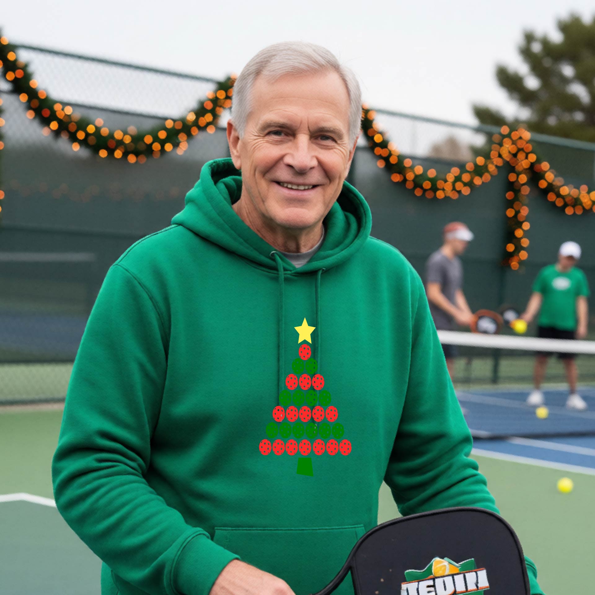A man wearing a green hoodie with a Christmas tree made of red and green pickleballs and a yellow star, in front of a pickleball court.