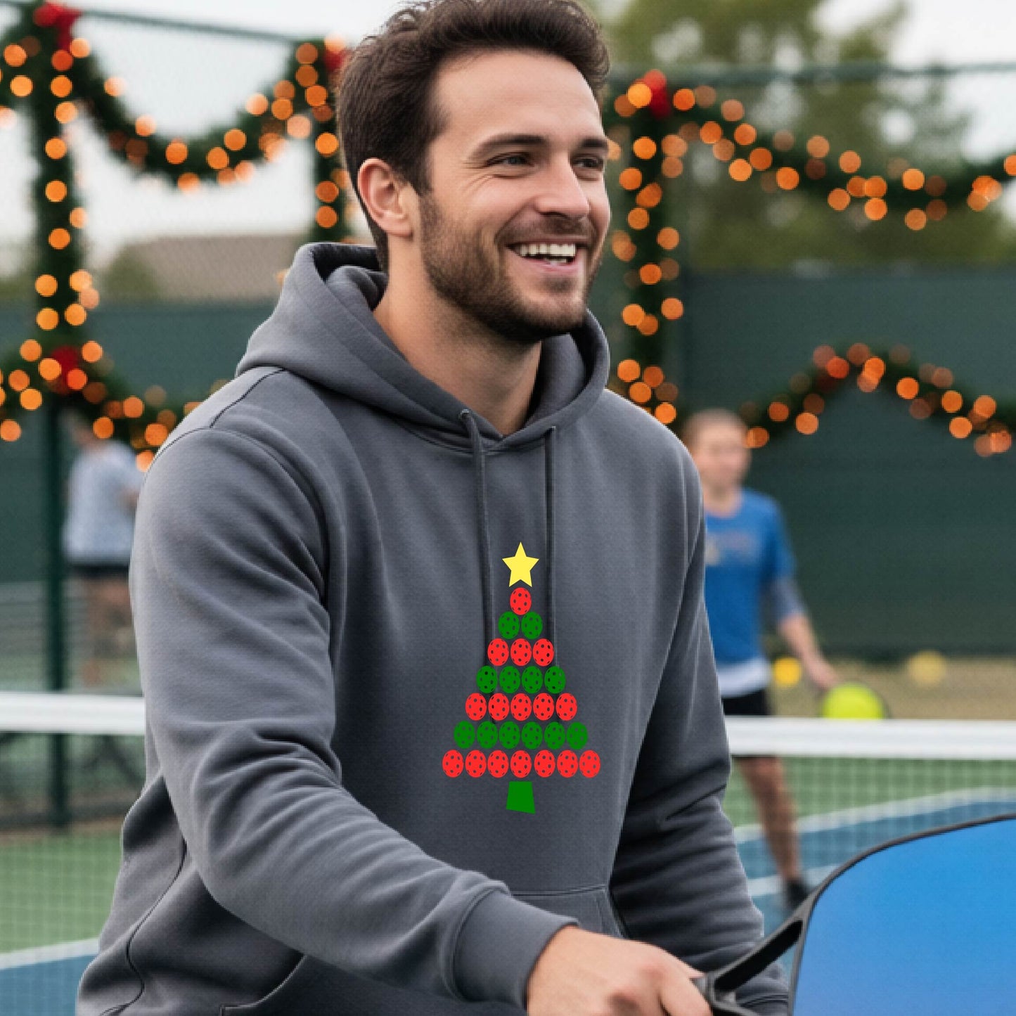 A man wearing a gray hoodie with a Christmas tree made of red and green pickleballs and a yellow star, in front of a pickleball court.