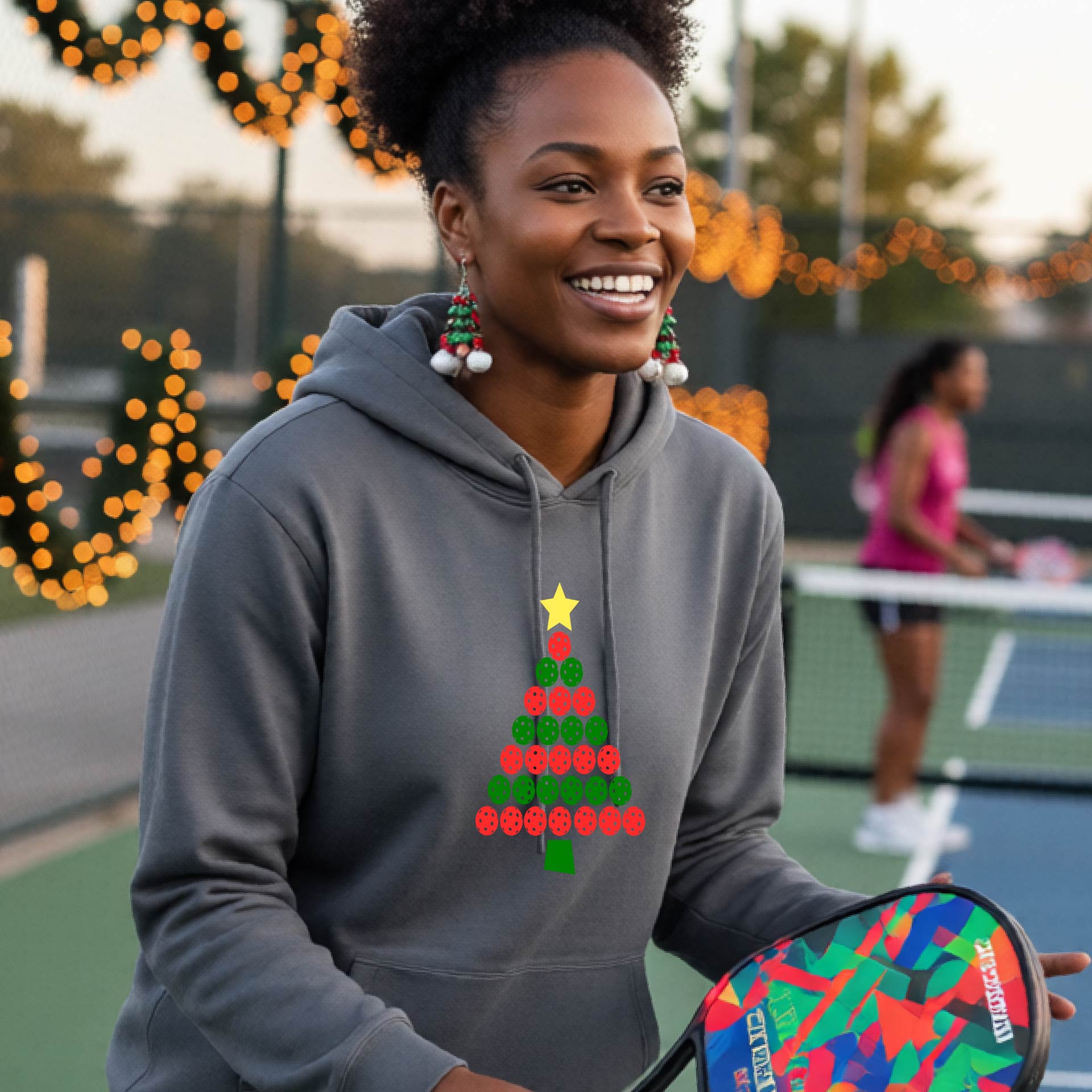 A woman wearing a gray hoodie with a Christmas tree made of red and green pickleballs and a yellow star, in front of a pickleball court.