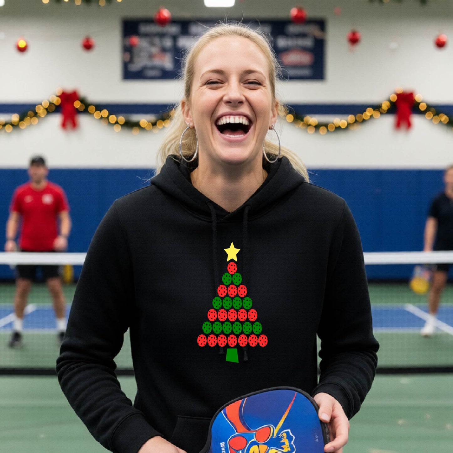 A woman viewed from behind wearing a black hoodie pullover with a Christmas tree made of red and green pickleballs and a yellow star on the back, set against a blurred indoor court decorated with holiday garlands.