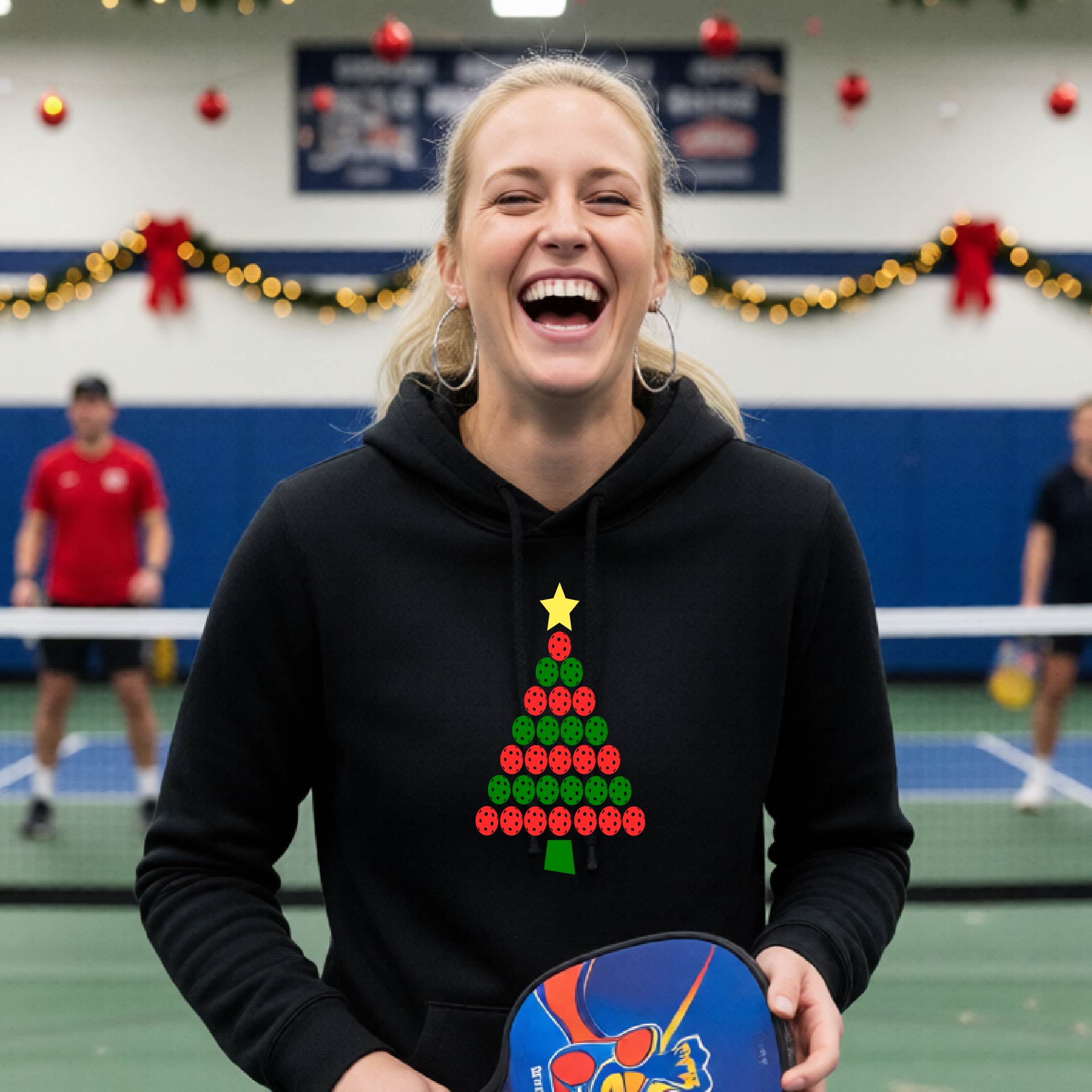 A woman viewed from behind wearing a black hoodie pullover with a Christmas tree made of red and green pickleballs and a yellow star on the back, set against a blurred indoor court decorated with holiday garlands.