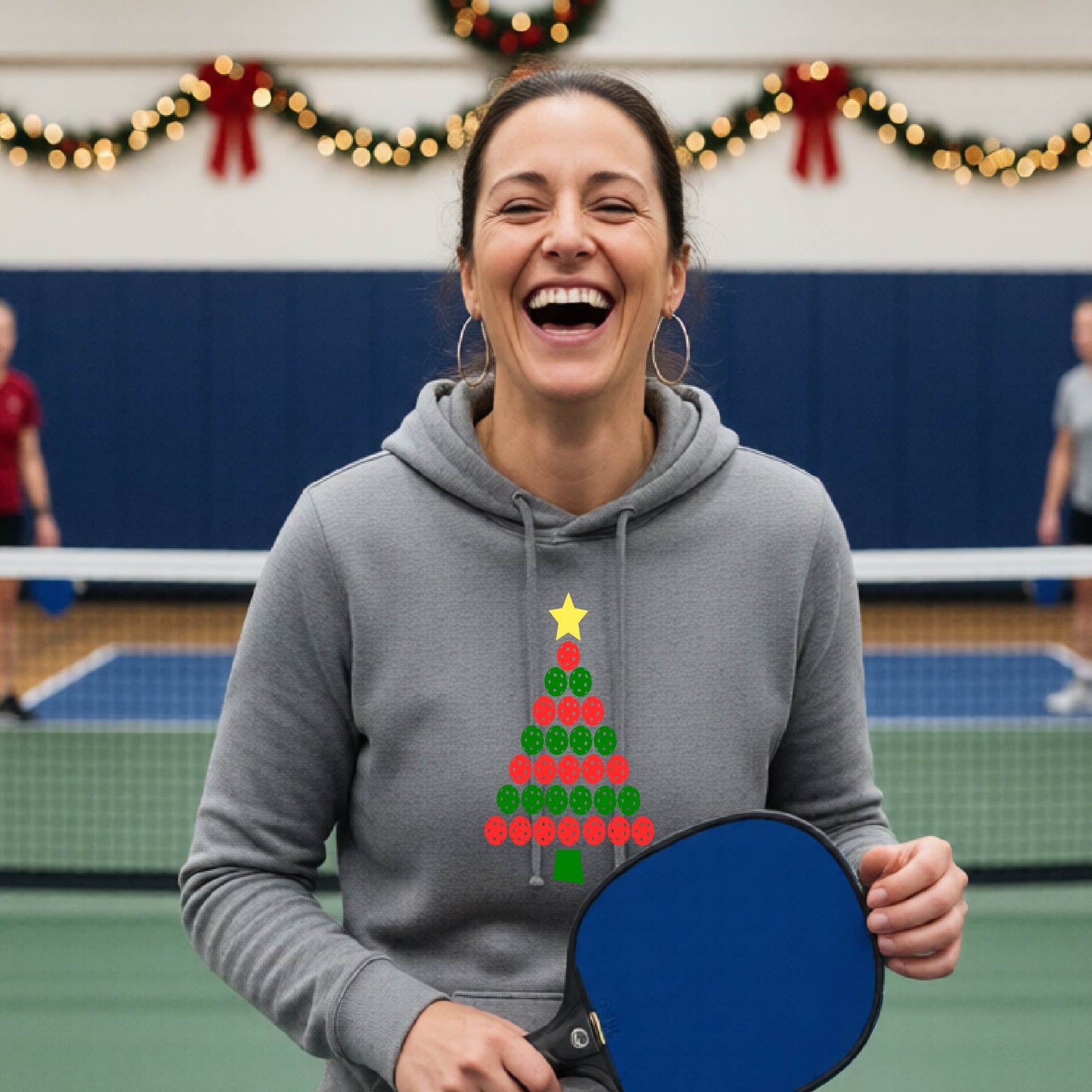 A woman viewed from behind wearing a gray hoodie pullover with a Christmas tree made of red and green pickleballs and a yellow star on the back, set against a blurred indoor court decorated with holiday garlands.