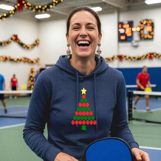 A woman viewed from behind wearing a navy hoodie pullover with a Christmas tree made of red and green pickleballs and a yellow star on the back, set against a blurred indoor court decorated with holiday garlands.