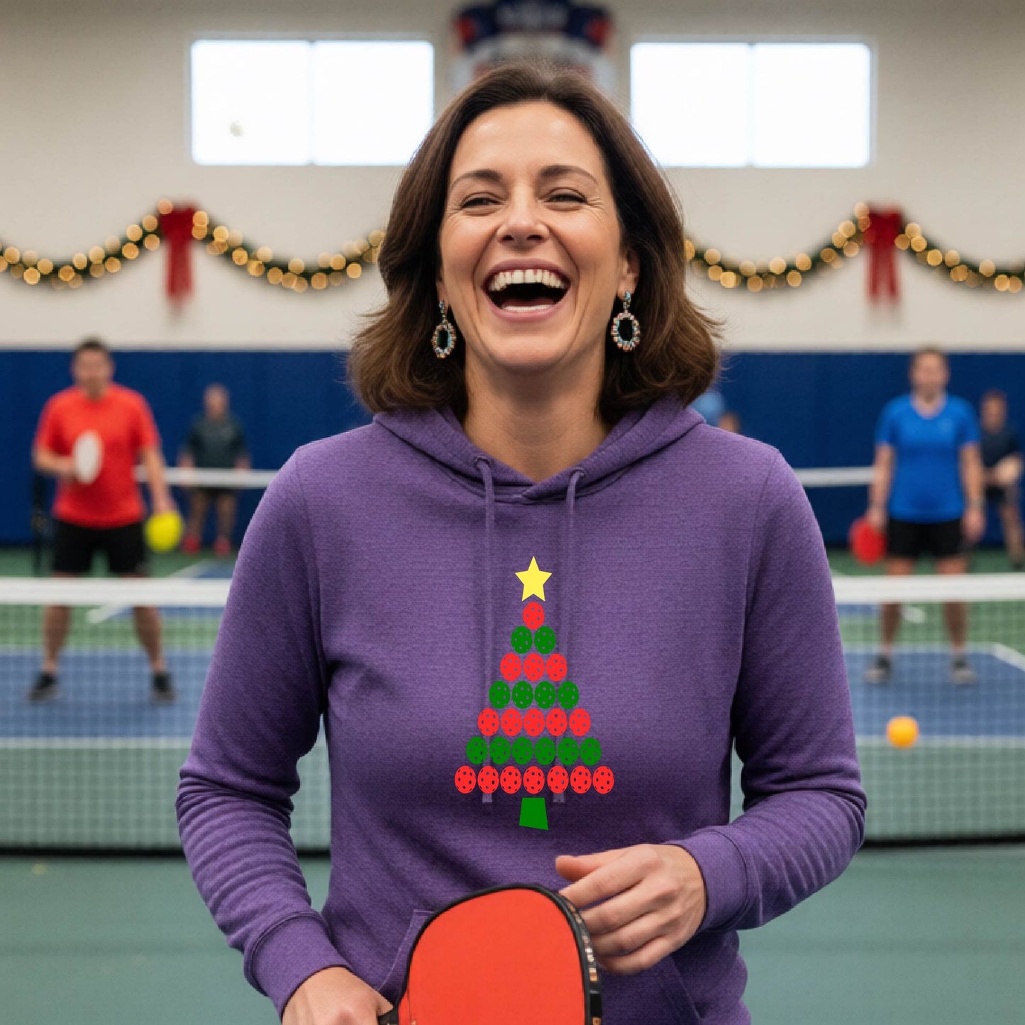 A woman viewed from behind wearing a purple hoodie pullover with a Christmas tree made of red and green pickleballs and a yellow star on the back, set against a blurred indoor court decorated with holiday garlands.