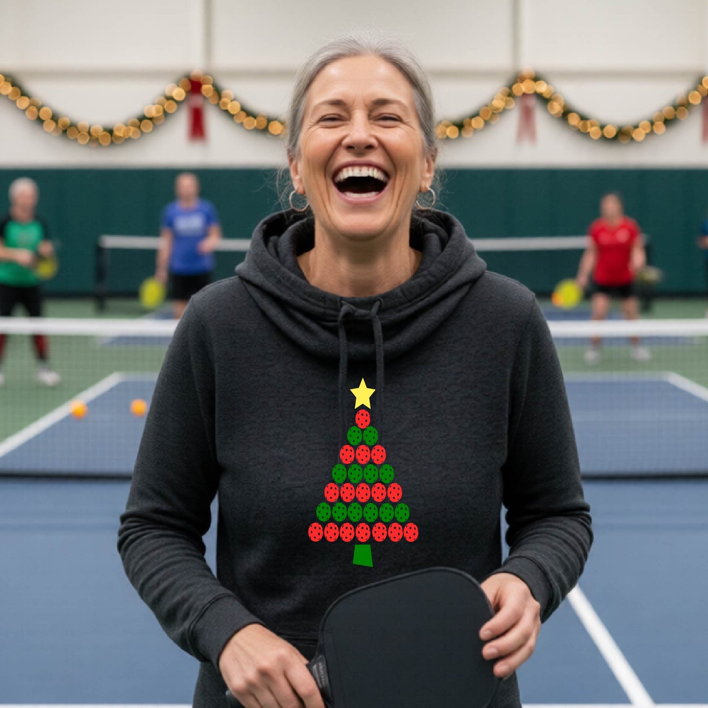 A black cowl-neck hooded sweatshirt featuring a pickleball graphic of a tree made of pickleballs and a Christmas themed background. 