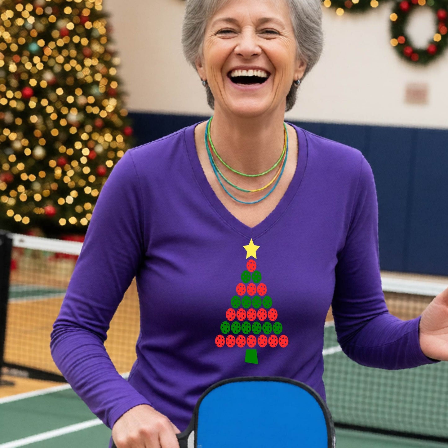 A smiling woman wearing a purple long-sleeve V-neck shirt with a Christmas tree made of red and green pickleballs and a yellow star, standing on a picleball court with a Christmas tree and holiday decorations in the background.