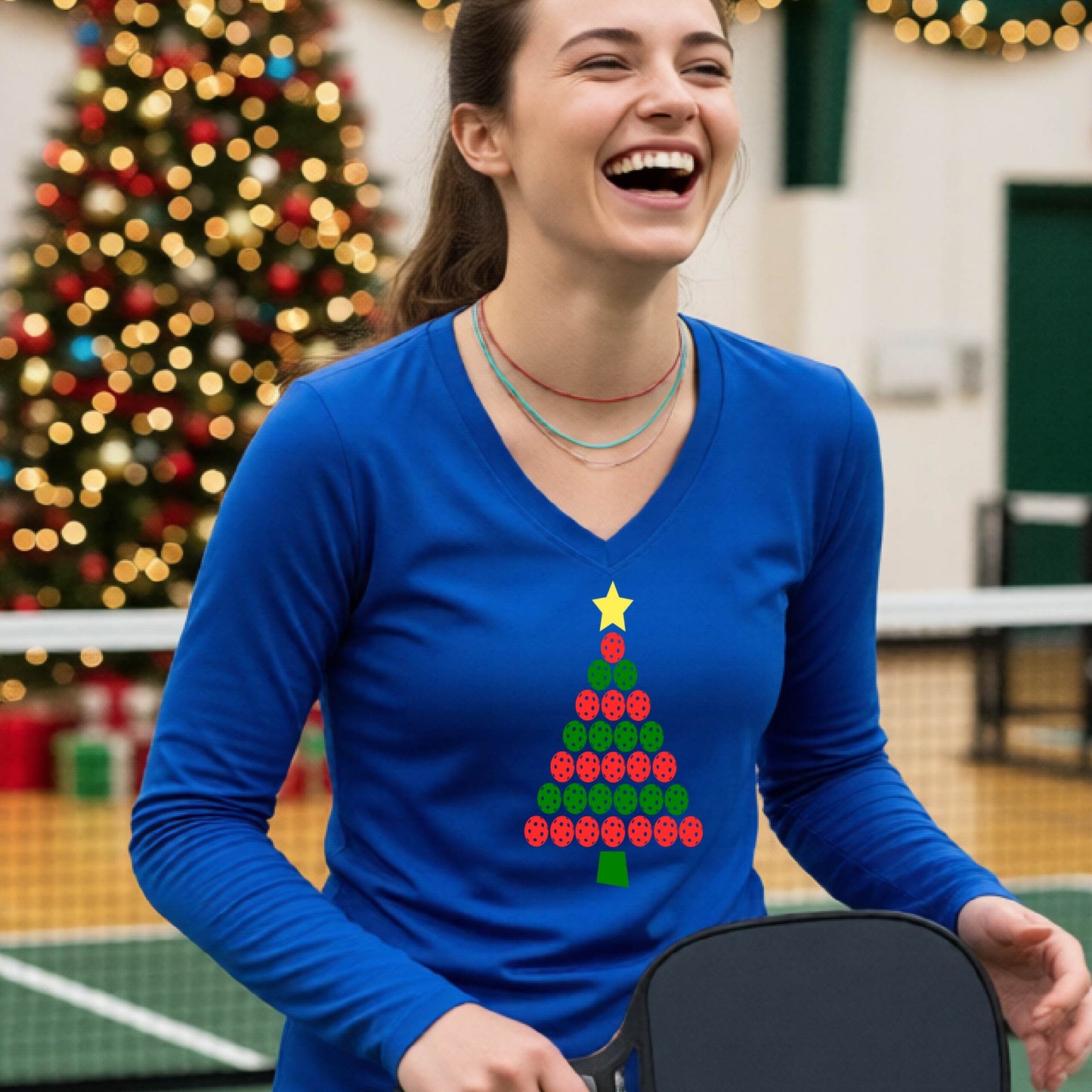 A smiling woman wearing a royal blue long-sleeve V-neck shirt with a Christmas tree made of red and green pickleballs and a yellow star, standing on a picleball court with a Christmas tree and holiday decorations in the background.