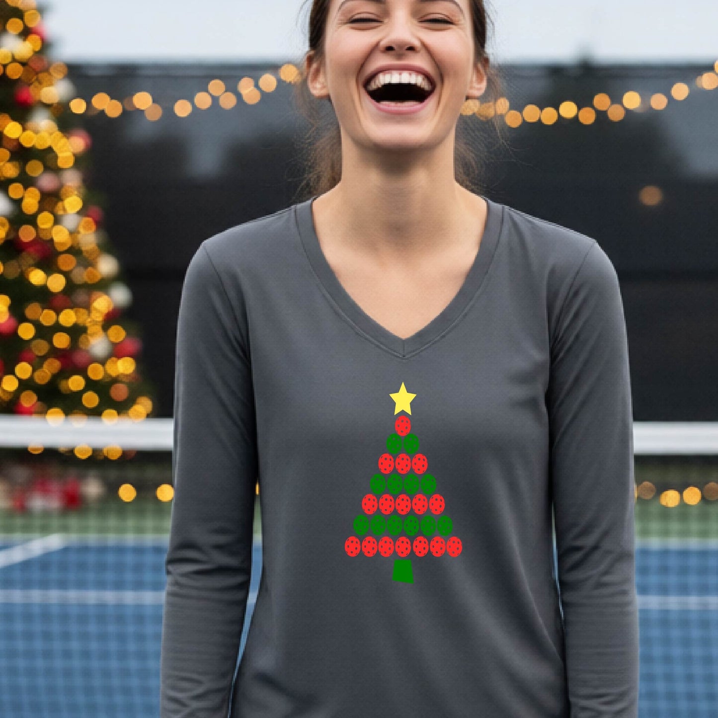 A smiling woman wearing a gray long-sleeve V-neck shirt with a Christmas tree made of red and green pickleballs and a yellow star, standing on a picleball court with a Christmas tree and holiday decorations in the background.