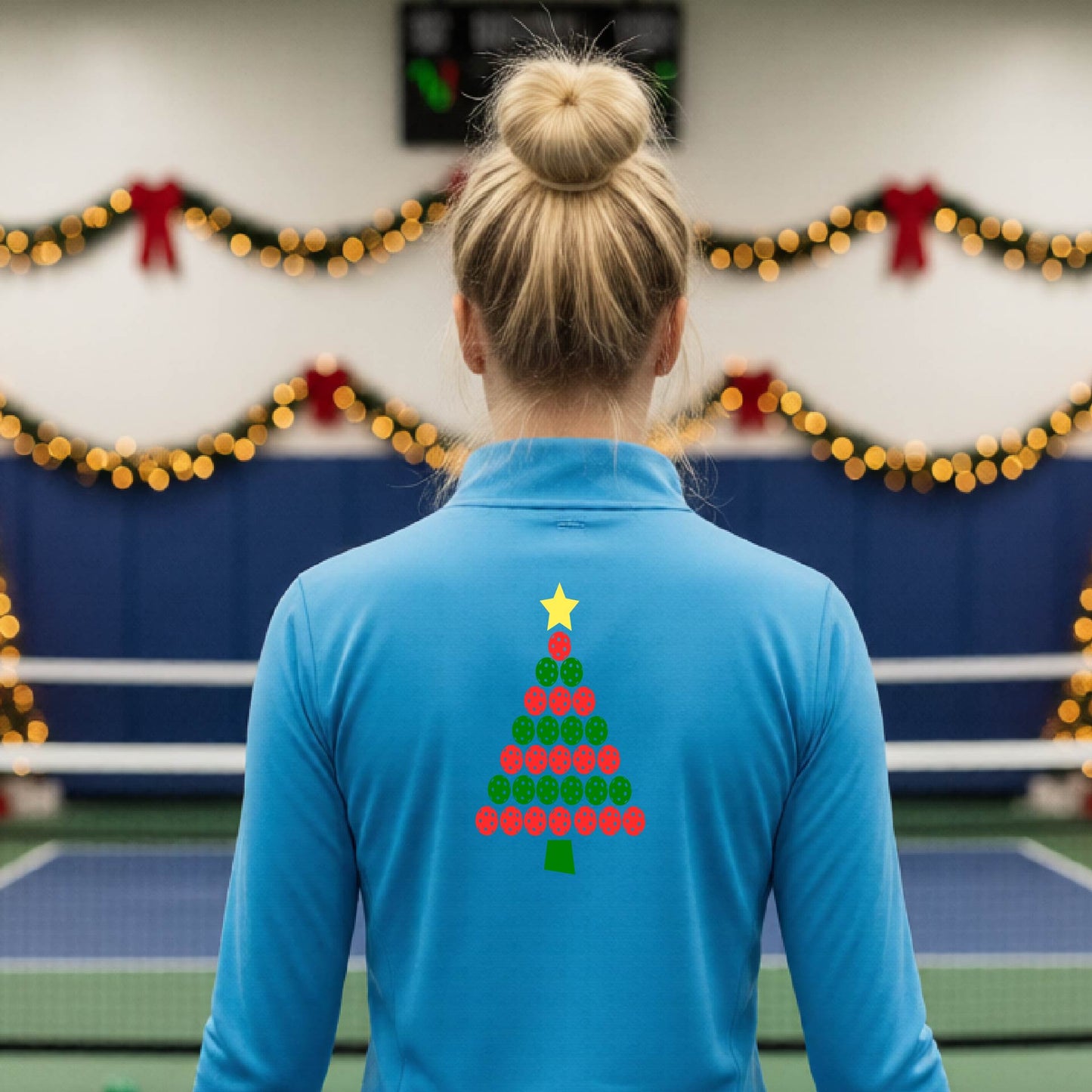 A woman viewed from behind wearing a bright blue athletic quarter-zip pullover with a Christmas tree made of red and green pickleballs and a yellow star on the back, set against a blurred indoor court decorated with holiday garlands.