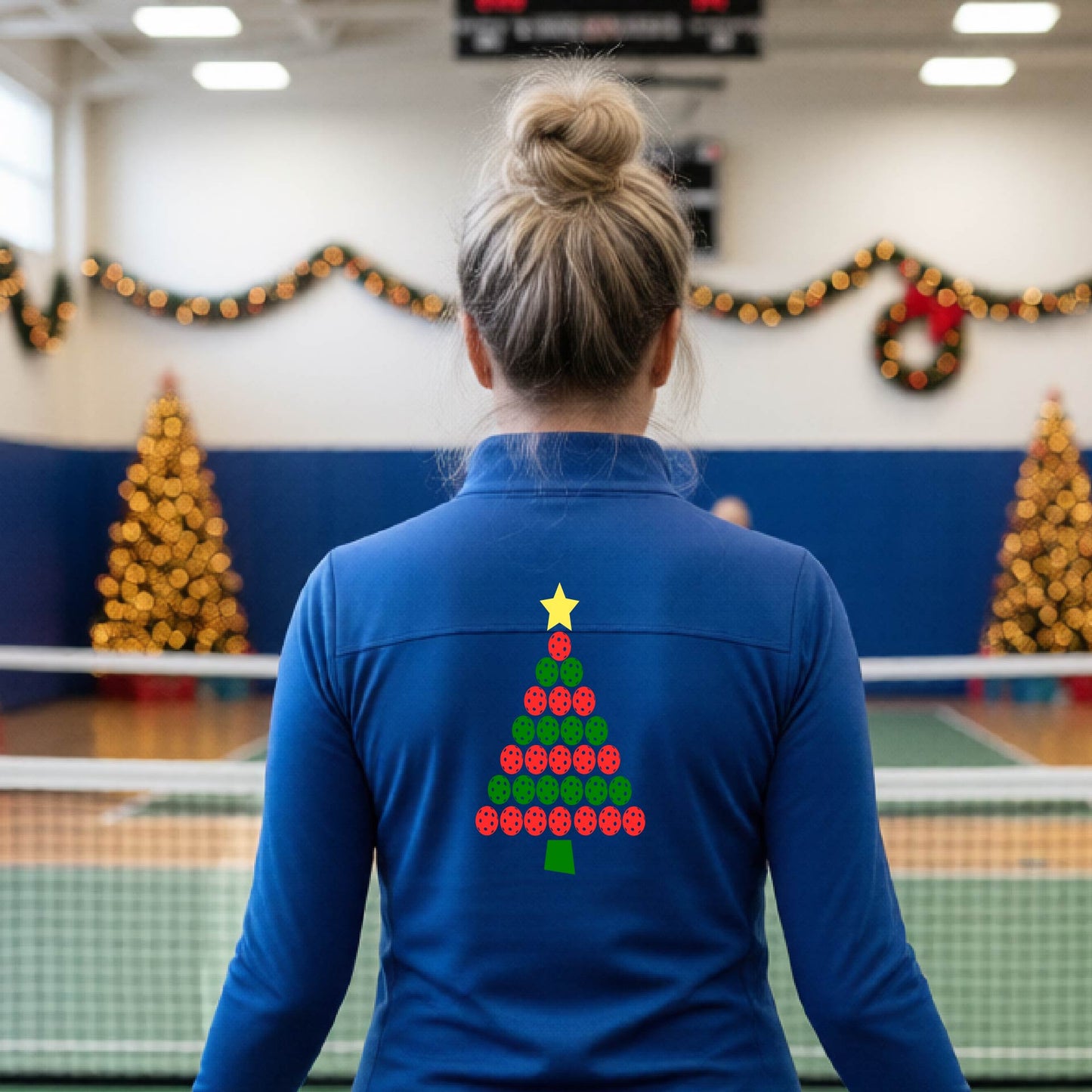 A woman viewed from behind wearing a royal blue athletic quarter-zip pullover with a Christmas tree made of red and green pickleballs and a yellow star on the back, set against a blurred indoor court decorated with holiday garlands.