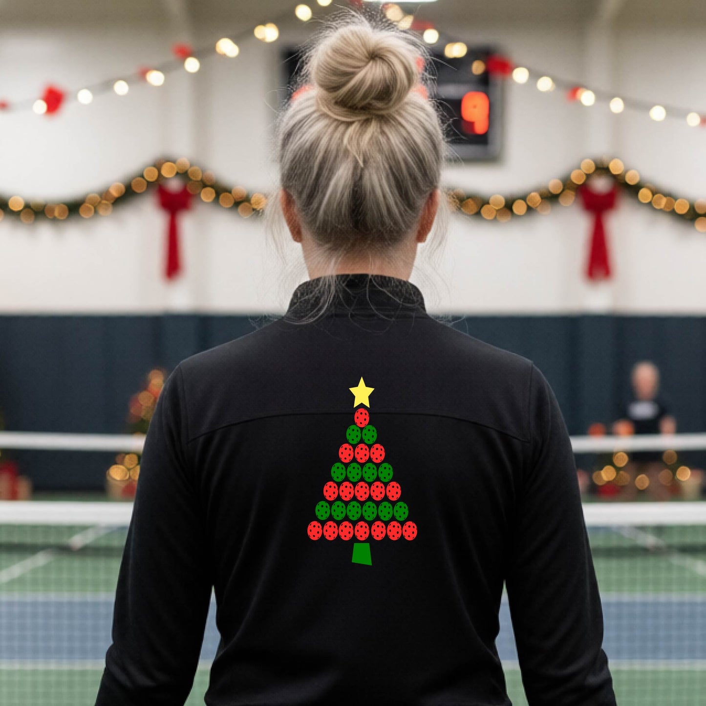 A woman viewed from behind wearing a black athletic quarter-zip pullover with a Christmas tree made of red and green pickleballs and a yellow star on the back, set against a blurred indoor court decorated with holiday garlands.