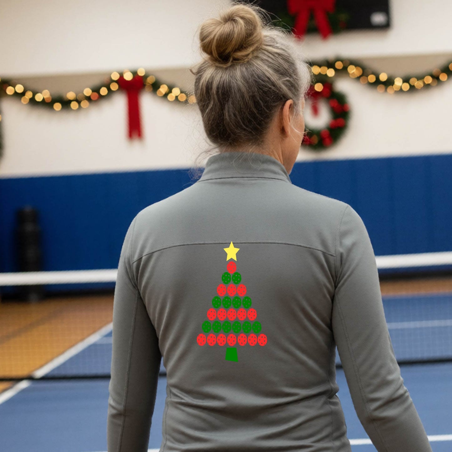 A woman viewed from behind wearing a gray athletic quarter-zip pullover with a Christmas tree made of red and green pickleballs and a yellow star on the back, set against a blurred indoor court decorated with holiday garlands.