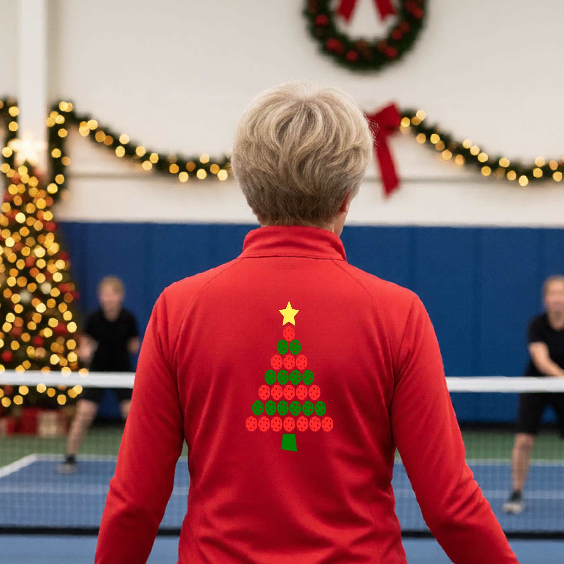 A woman viewed from behind wearing a bright red athletic quarter-zip pullover with a Christmas tree made of red and green pickleballs and a yellow star on the back, set against a blurred indoor court decorated with holiday garlands.