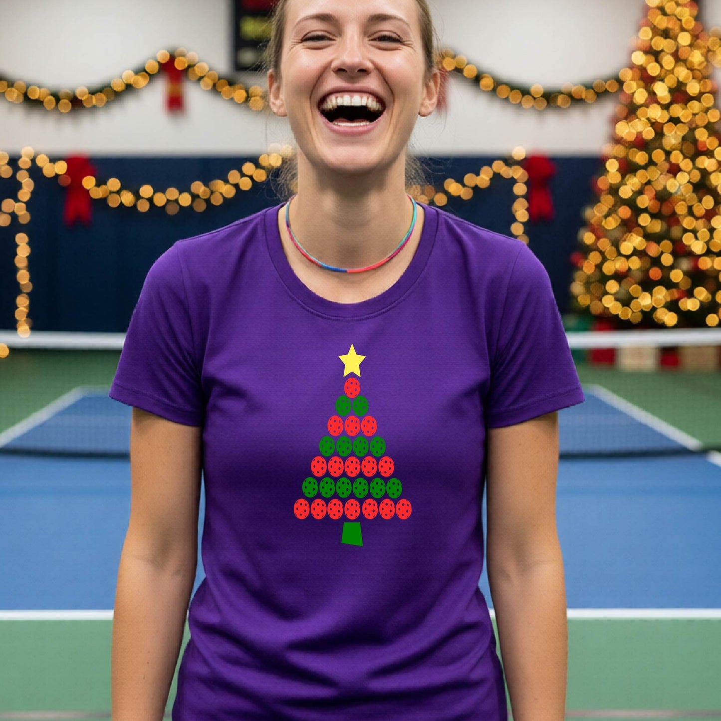 A woman smiling while wearing a purple t-shirt with a Christmas tree graphic made of red and green pickleballs and a yellow star, set against a festive indoor background with a blurred Christmas tree and lights.