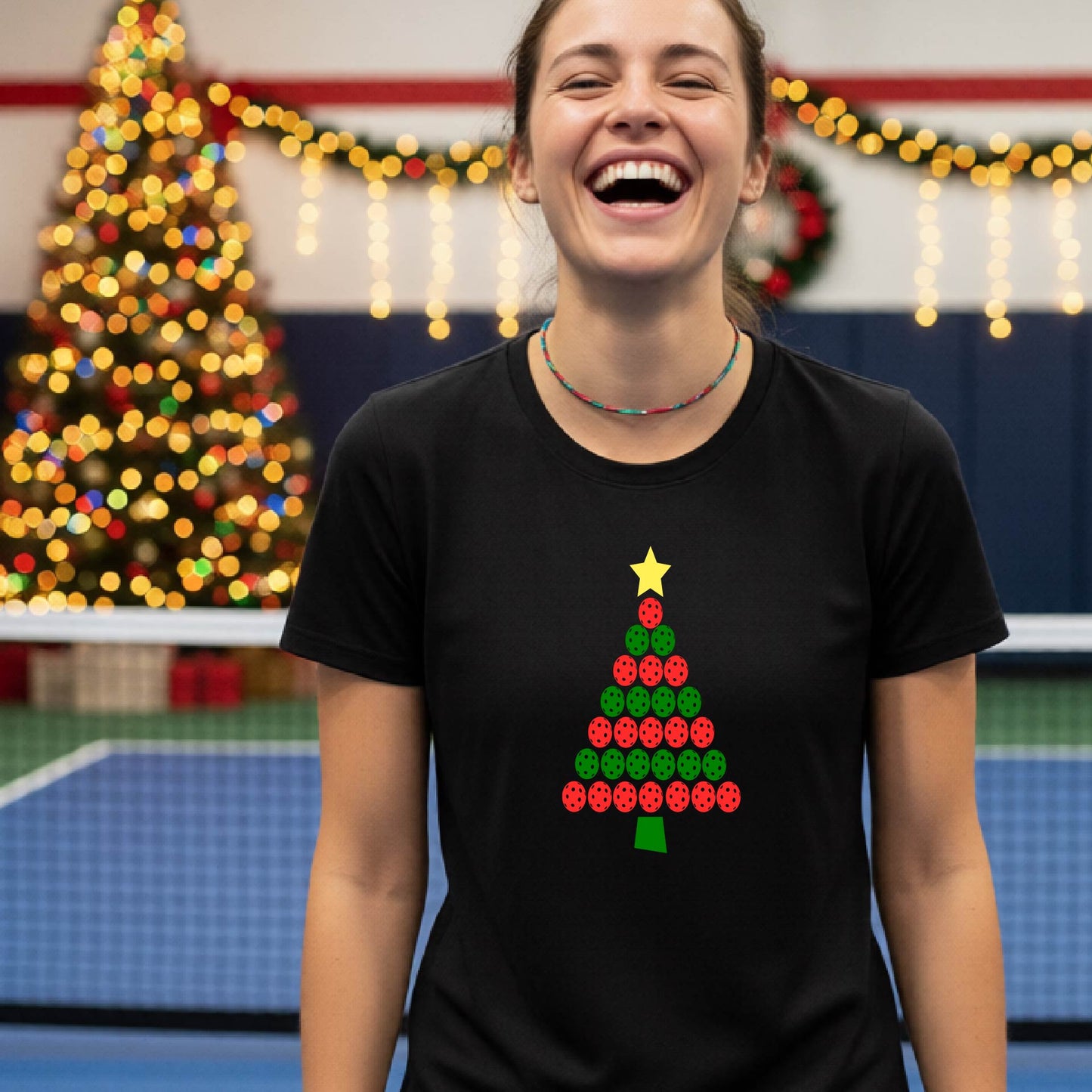 A woman smiling while wearing a black t-shirt with a Christmas tree graphic made of red and green pickleballs and a yellow star, set against a festive indoor background with a blurred Christmas tree and lights.