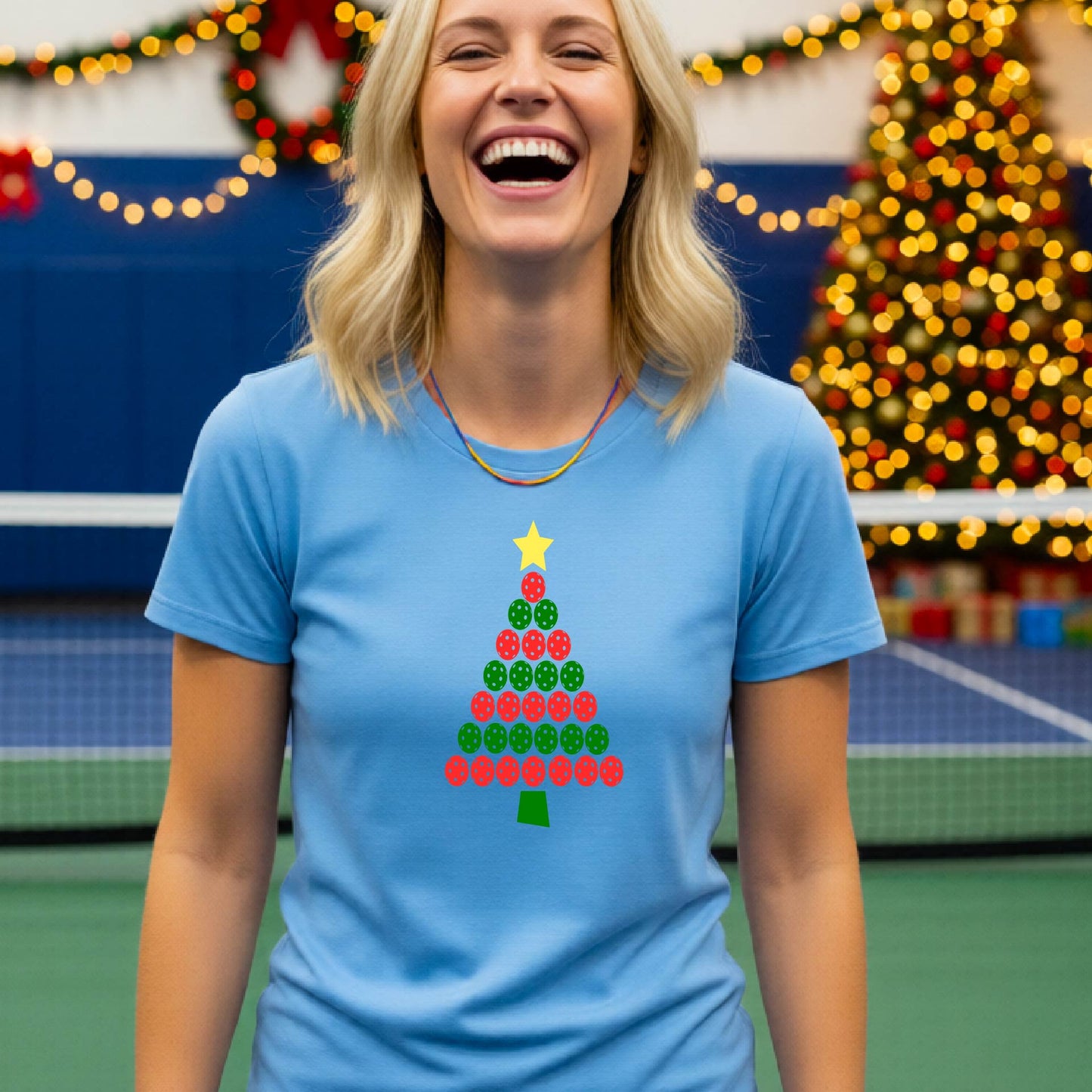 A woman smiling while wearing a light blue t-shirt with a Christmas tree graphic made of red and green pickleballs and a yellow star, set against a festive indoor background with a blurred Christmas tree and lights.