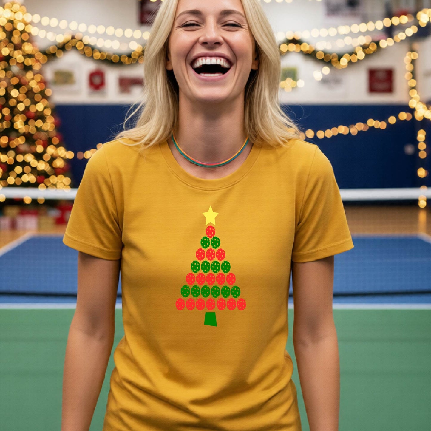 A woman smiling while wearing a mustard yellow gold t-shirt with a Christmas tree graphic made of red and green pickleballs and a yellow star, set against a festive indoor background with a blurred Christmas tree and lights.