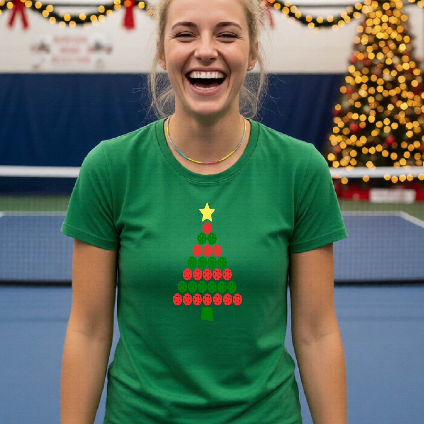 A woman smiling while wearing a green t-shirt with a Christmas tree graphic made of red and green pickleballs and a yellow star, set against a festive indoor background with a blurred Christmas tree and lights.