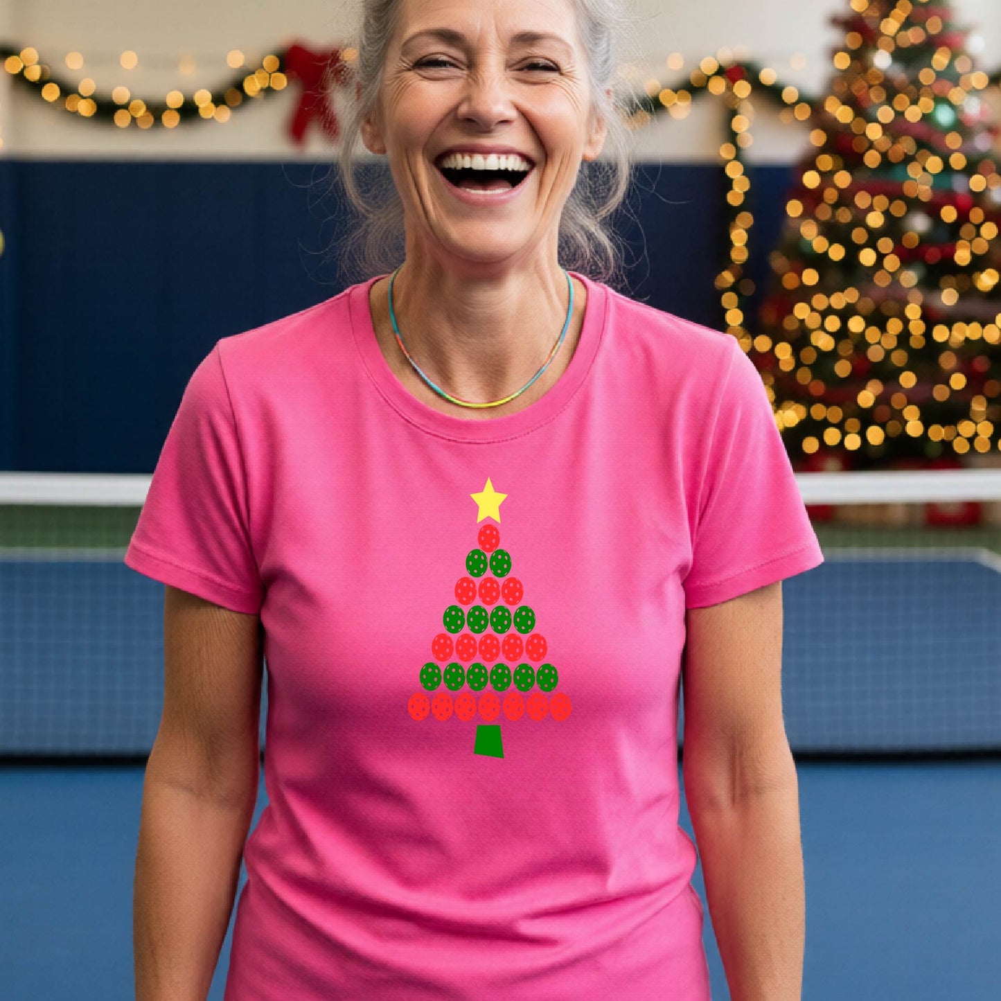A woman smiling while wearing a bright pink t-shirt with a Christmas tree graphic made of red and green pickleballs and a yellow star, set against a festive indoor background with a blurred Christmas tree and lights.