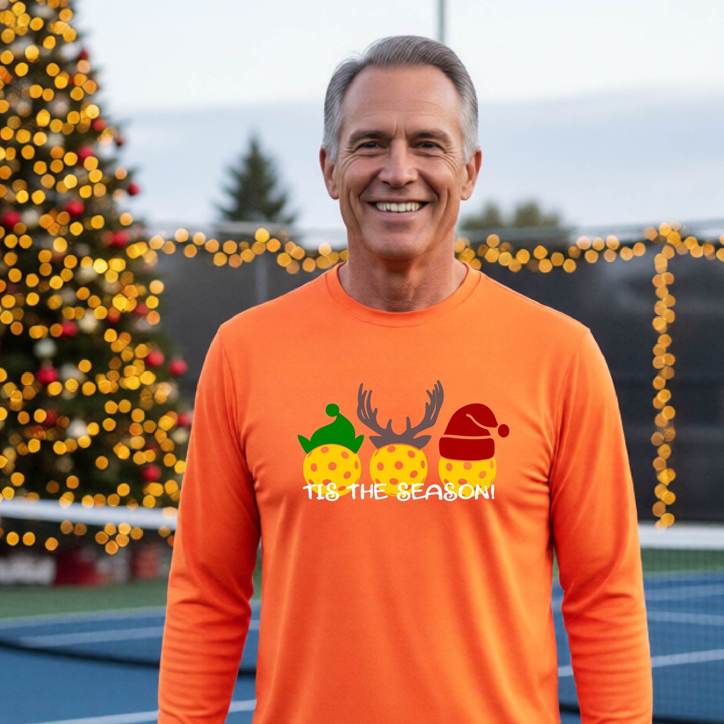 A man wearing an orange graphic long-sleeve t-shirt with a Christmas-themed design of three pickleballs wearing holiday hats and the text 'TIS THE SEASON!'. The background shows a festive blurred Christmas tree and a pickleball court.