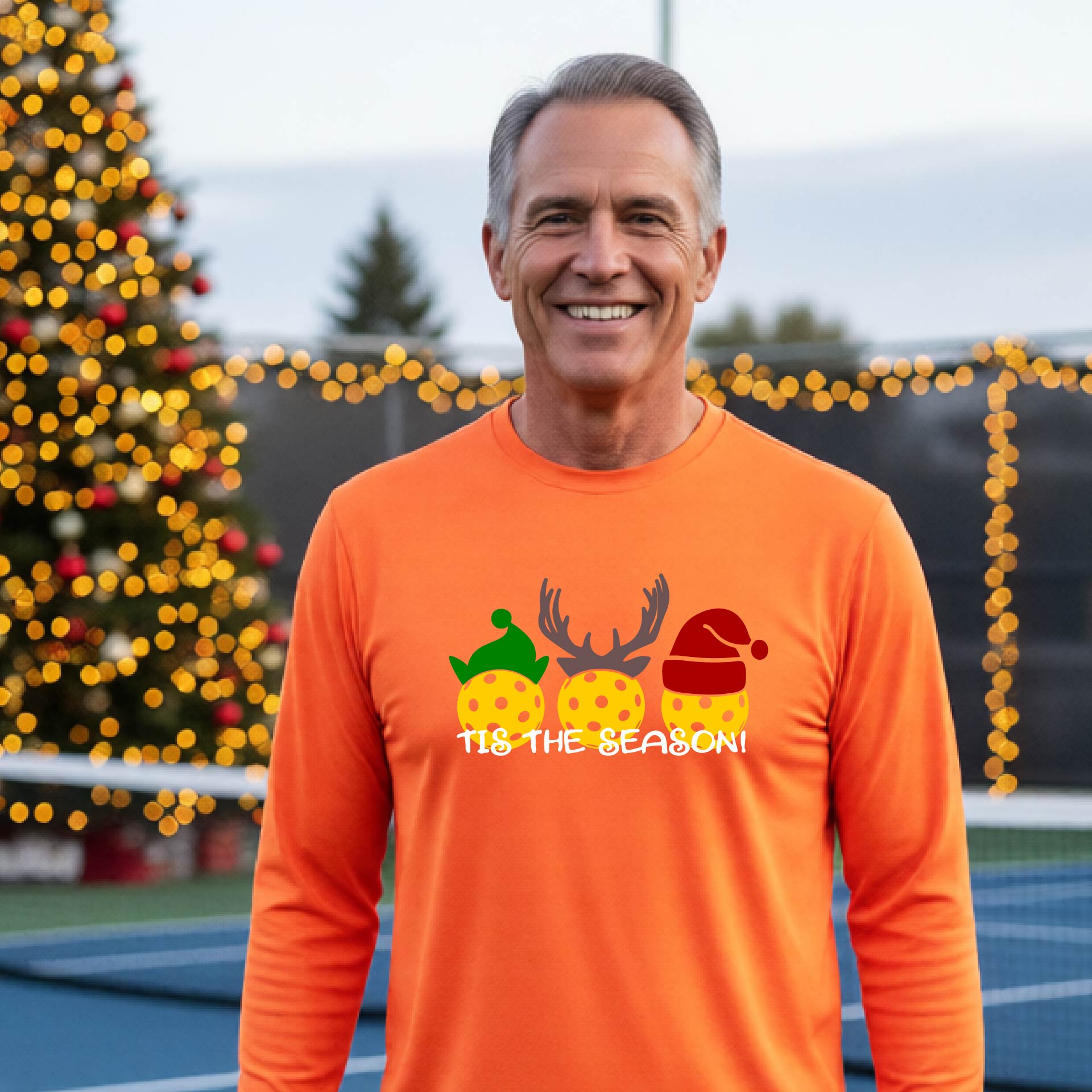 A man wearing an orange graphic long-sleeve t-shirt with a Christmas-themed design of three pickleballs wearing holiday hats and the text 'TIS THE SEASON!'. The background shows a festive blurred Christmas tree and a pickleball court.