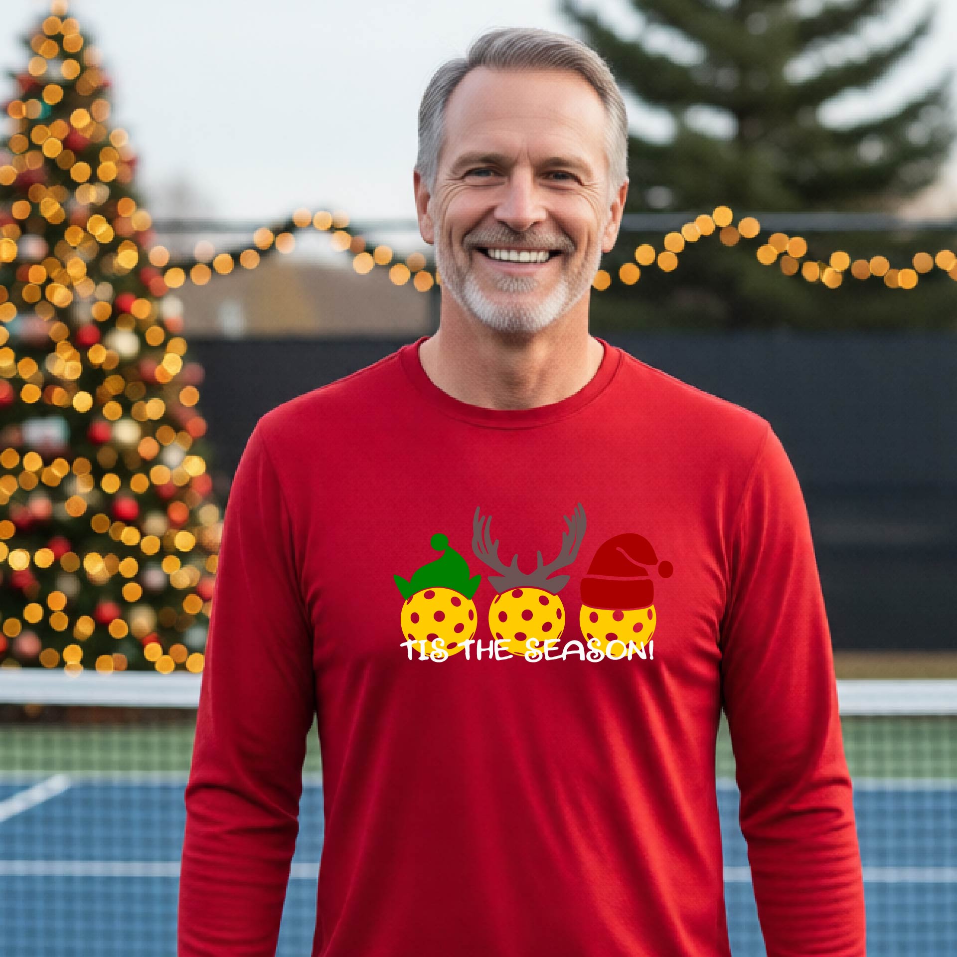 A man wearing a red graphic long-sleeve t-shirt with a Christmas-themed design of three pickleballs wearing holiday hats and the text 'TIS THE SEASON!'. The background shows a festive blurred Christmas tree and a pickleball court.