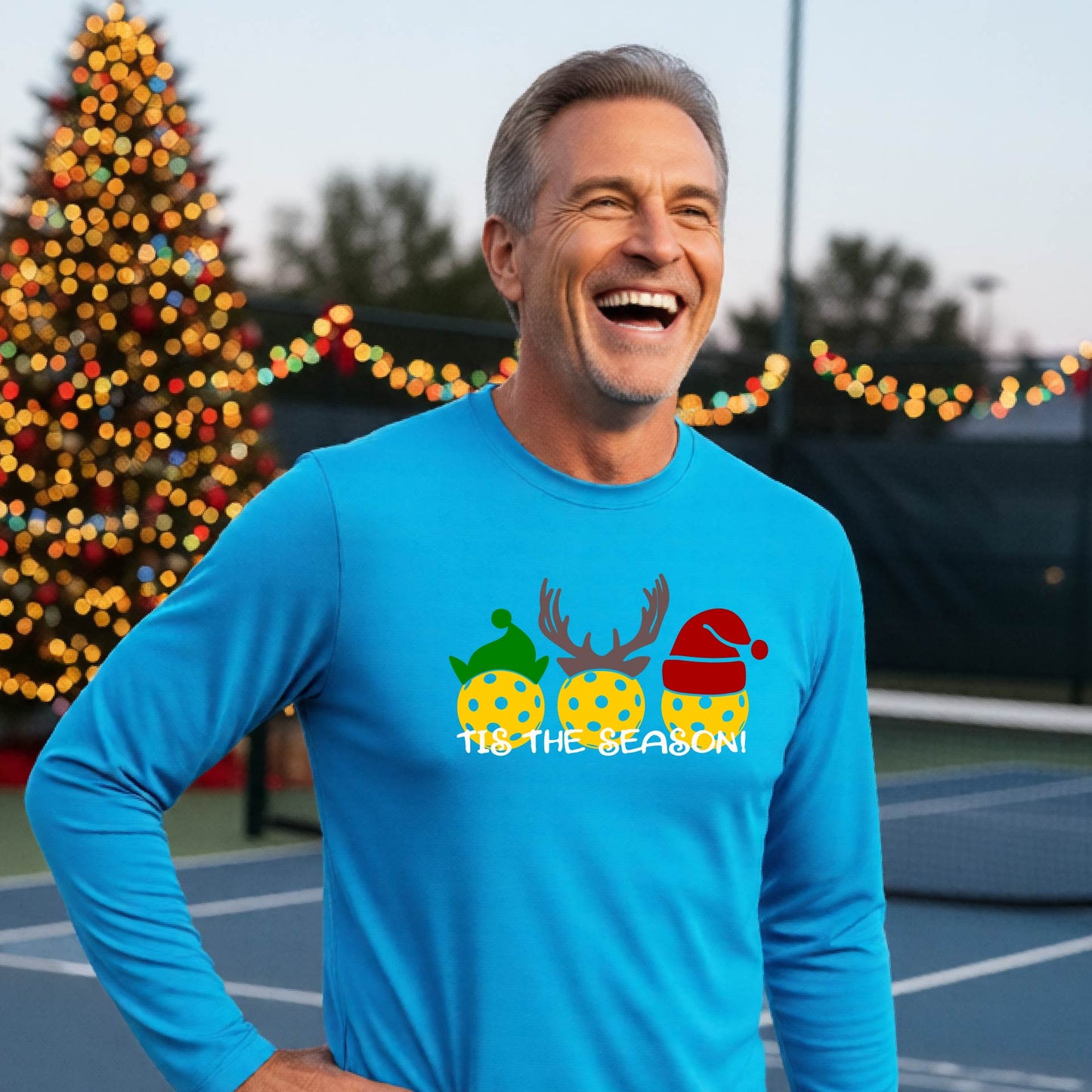 A man wearing a bright blue graphic long-sleeve t-shirt with a Christmas-themed design of three pickleballs wearing holiday hats and the text 'TIS THE SEASON!'. The background shows a festive blurred Christmas tree and a pickleball court.