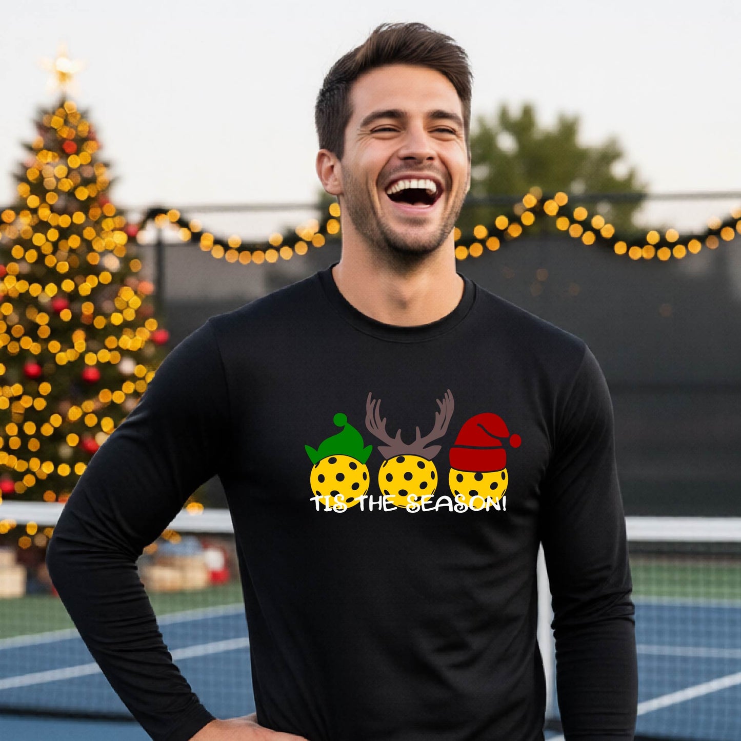 A man wearing a black graphic long-sleeve t-shirt with a Christmas-themed design of three pickleballs wearing holiday hats and the text 'TIS THE SEASON!'. The background shows a festive blurred Christmas tree and a pickleball court.