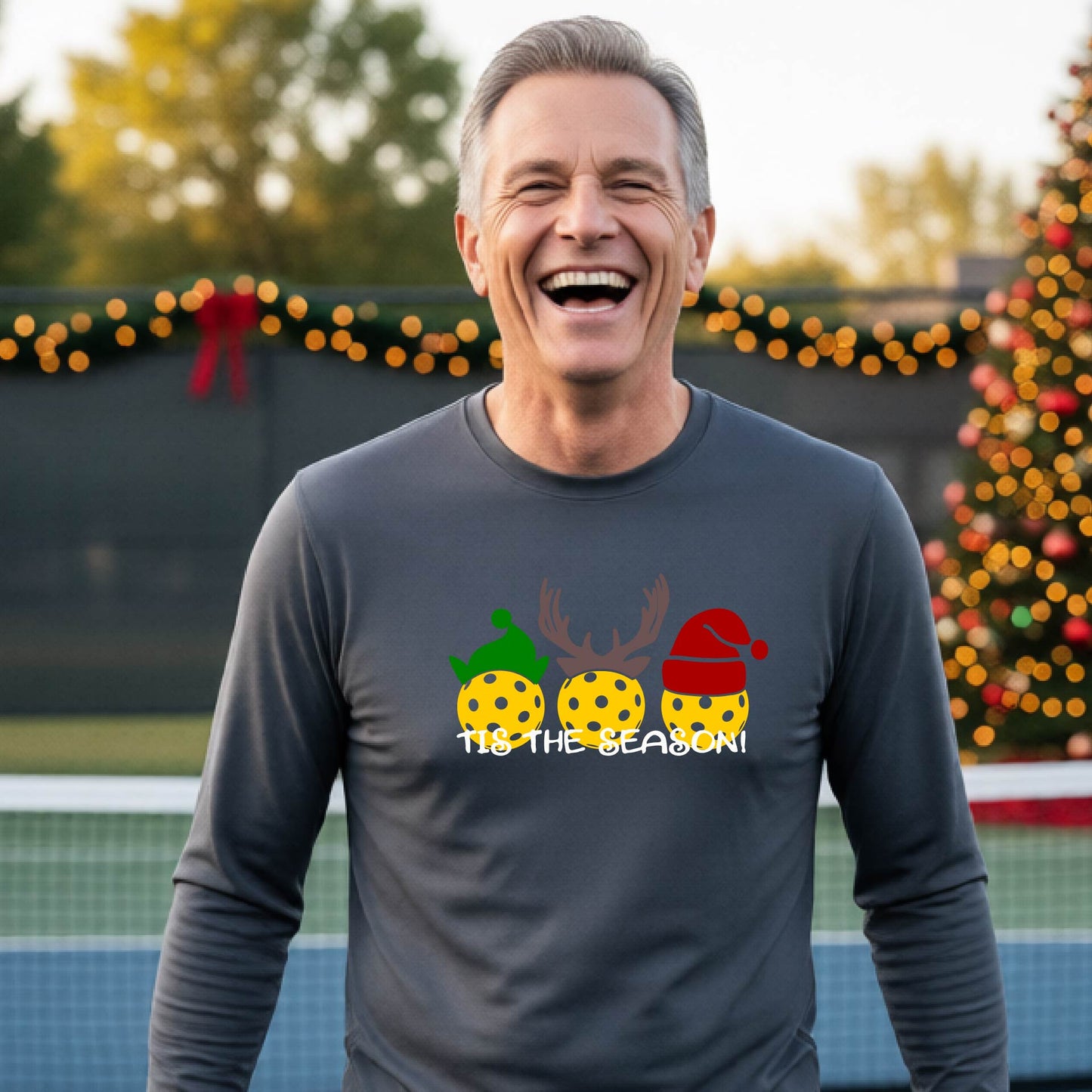 A man wearing a gray graphic long-sleeve t-shirt with a Christmas-themed design of three pickleballs wearing holiday hats and the text 'TIS THE SEASON!'. The background shows a festive blurred Christmas tree and a pickleball court.